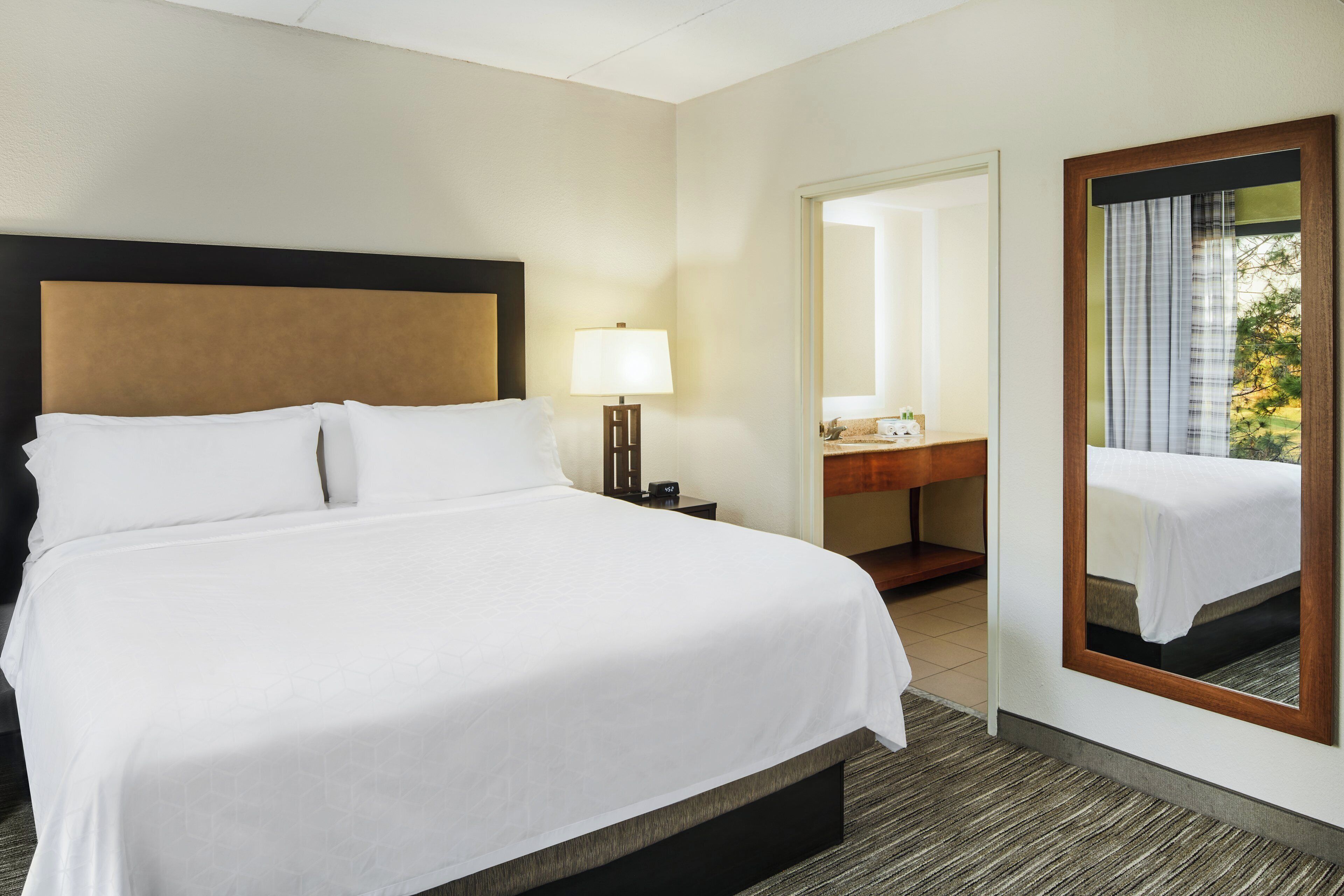 A hotel room with a large bed, a bedside lamp, a mirror, a small table with toiletries, and a view of trees through the window.