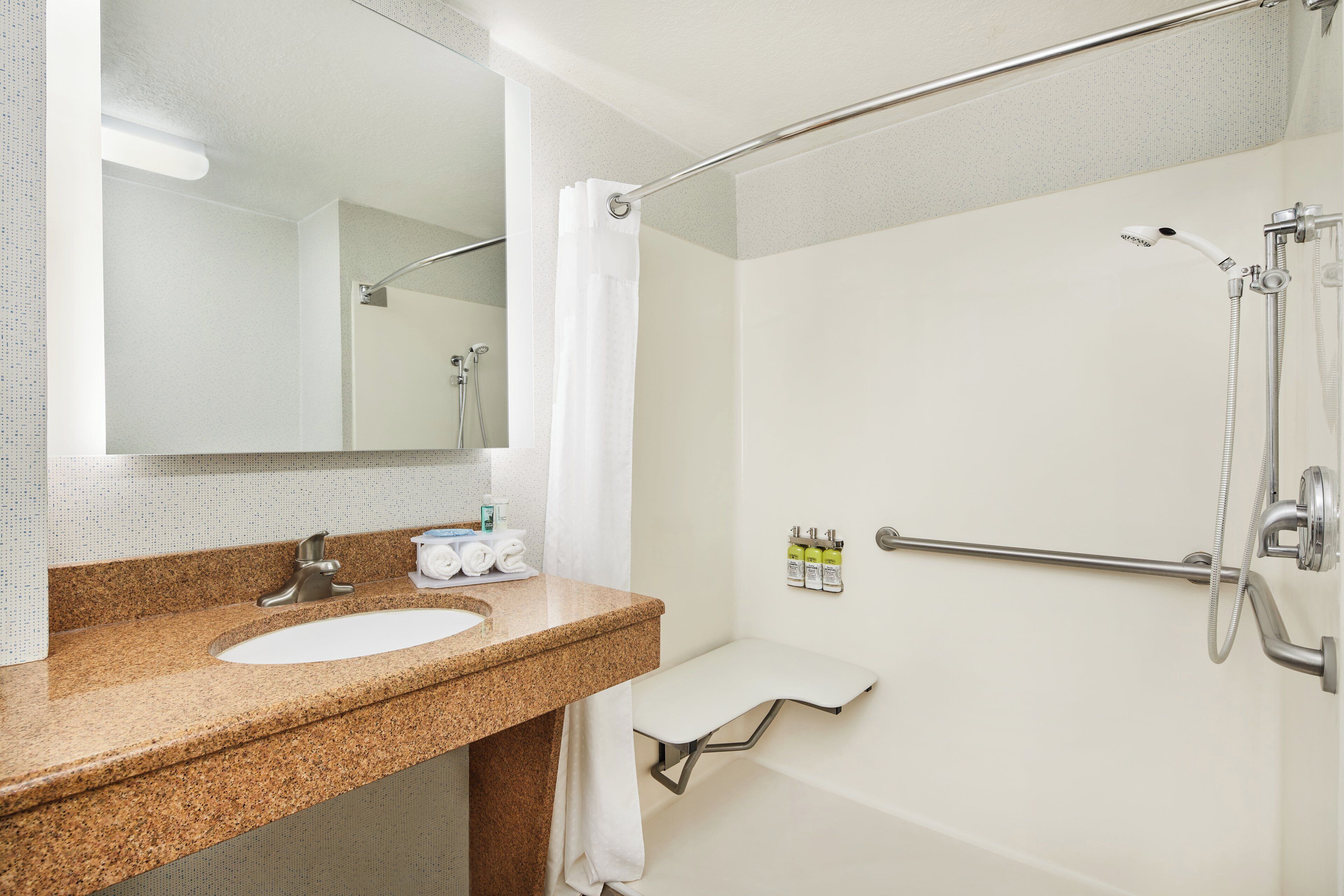 Un baño con lavamanos de encimera de granito, un espejo, ducha con cabezal de ducha manual y un perchero para toallas.