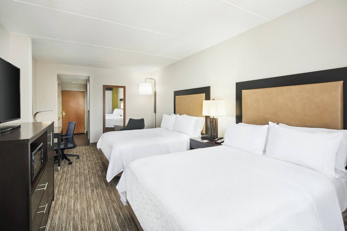 Une chambre d’hôtel avec deux lits, un bureau avec une télévision, une chaise, une lampe et une porte donnant sur une autre pièce.