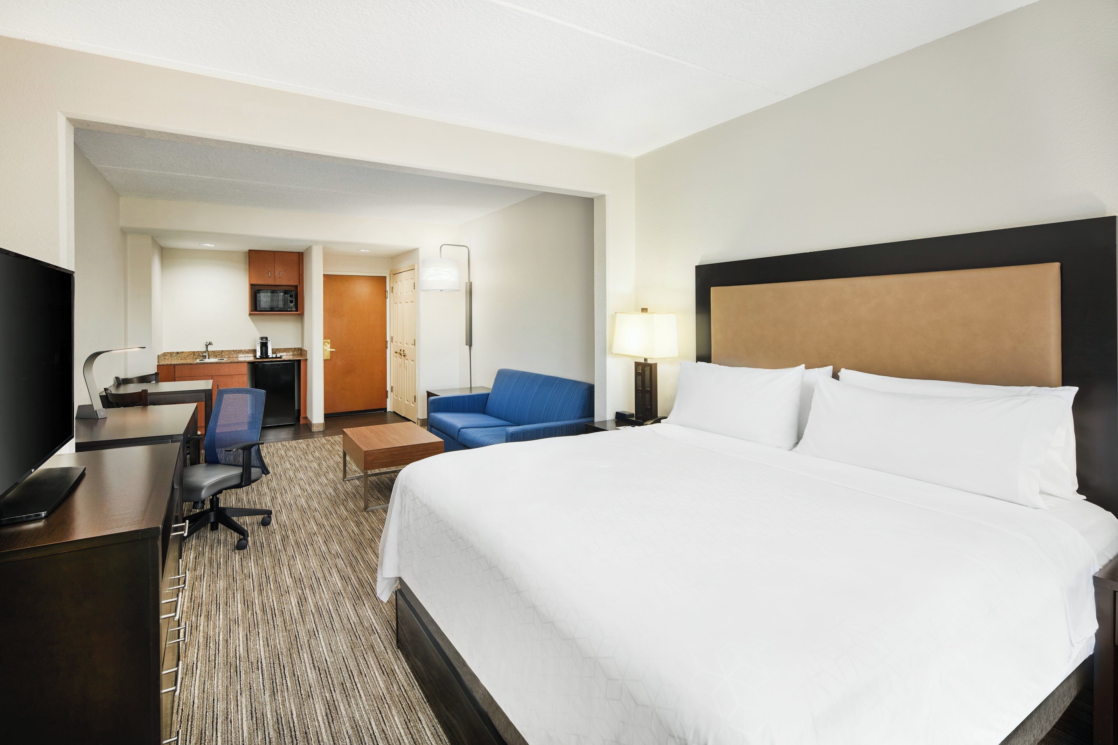 A hotel room with a large bed, a desk with a computer, a chair, a sofa, and a kitchenette.