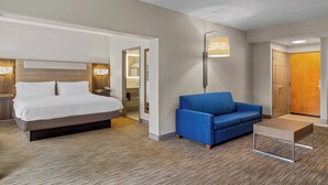 A modern hotel room with a large bed, a blue sofa, a small table, and a wall-mounted lamp. - Holiday Inn Express & Suites Jacksonville SE- Med Ctr Area by IHG (Jacksonville)