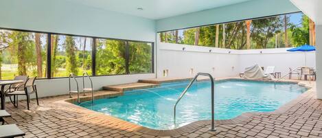 An indoor swimming pool with clear views of trees outside, a tiled floor, and a covered seating area.