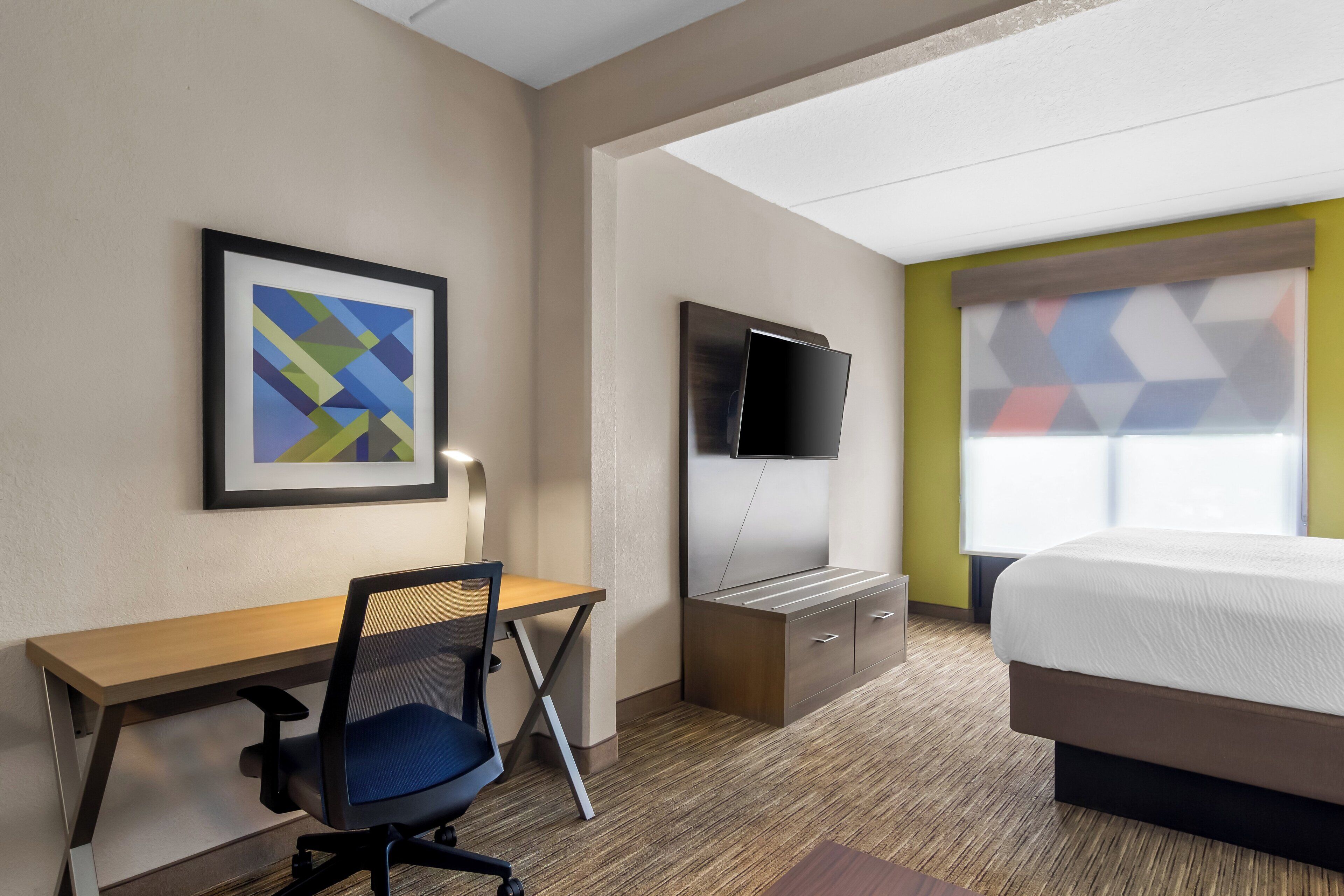 Habitación de hotel con cama, escritorio, silla, televisor y un cuadro en la pared.