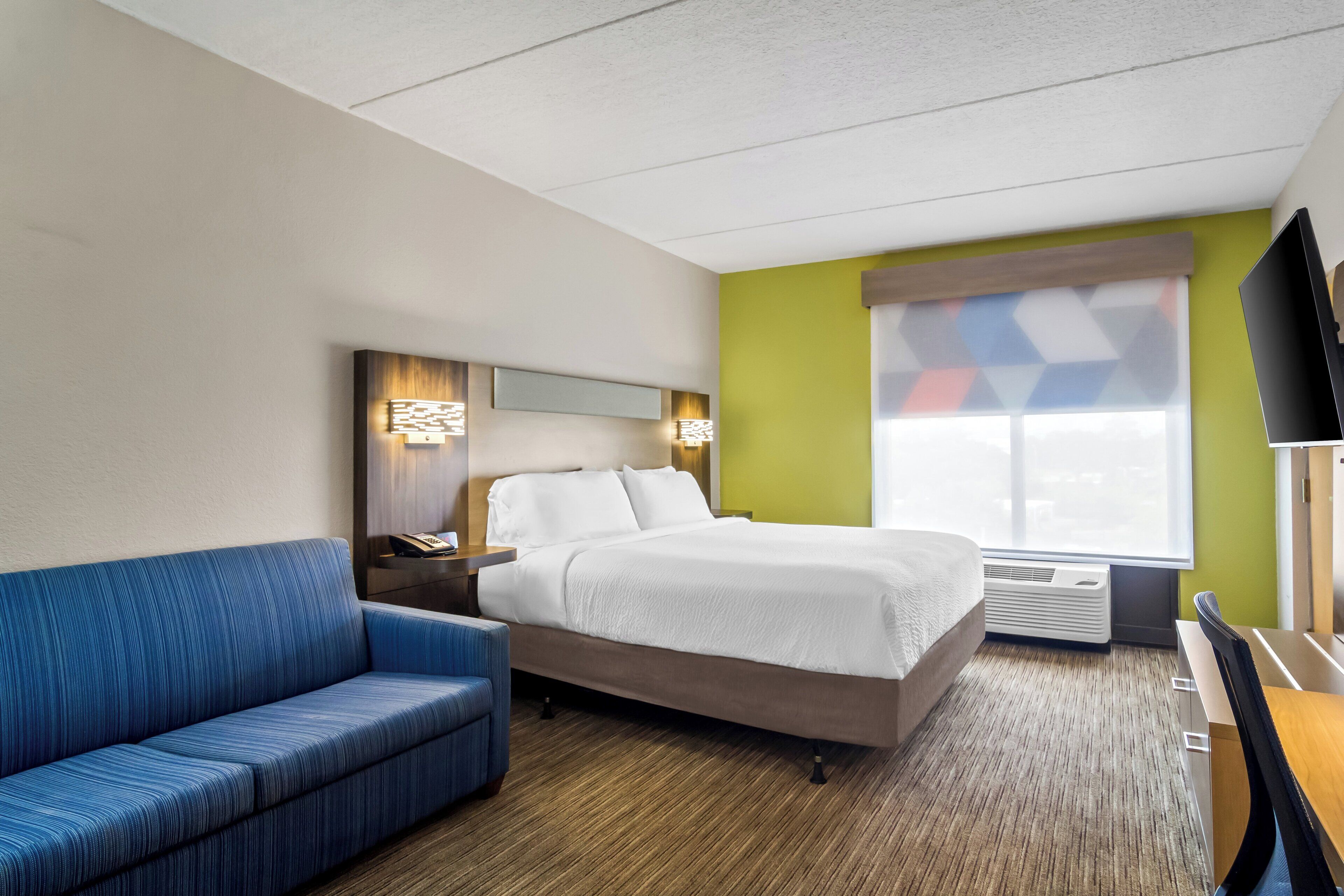 A hotel room with a bed, a blue sofa, a desk with a chair, a TV, and a window with a geometric curtain.