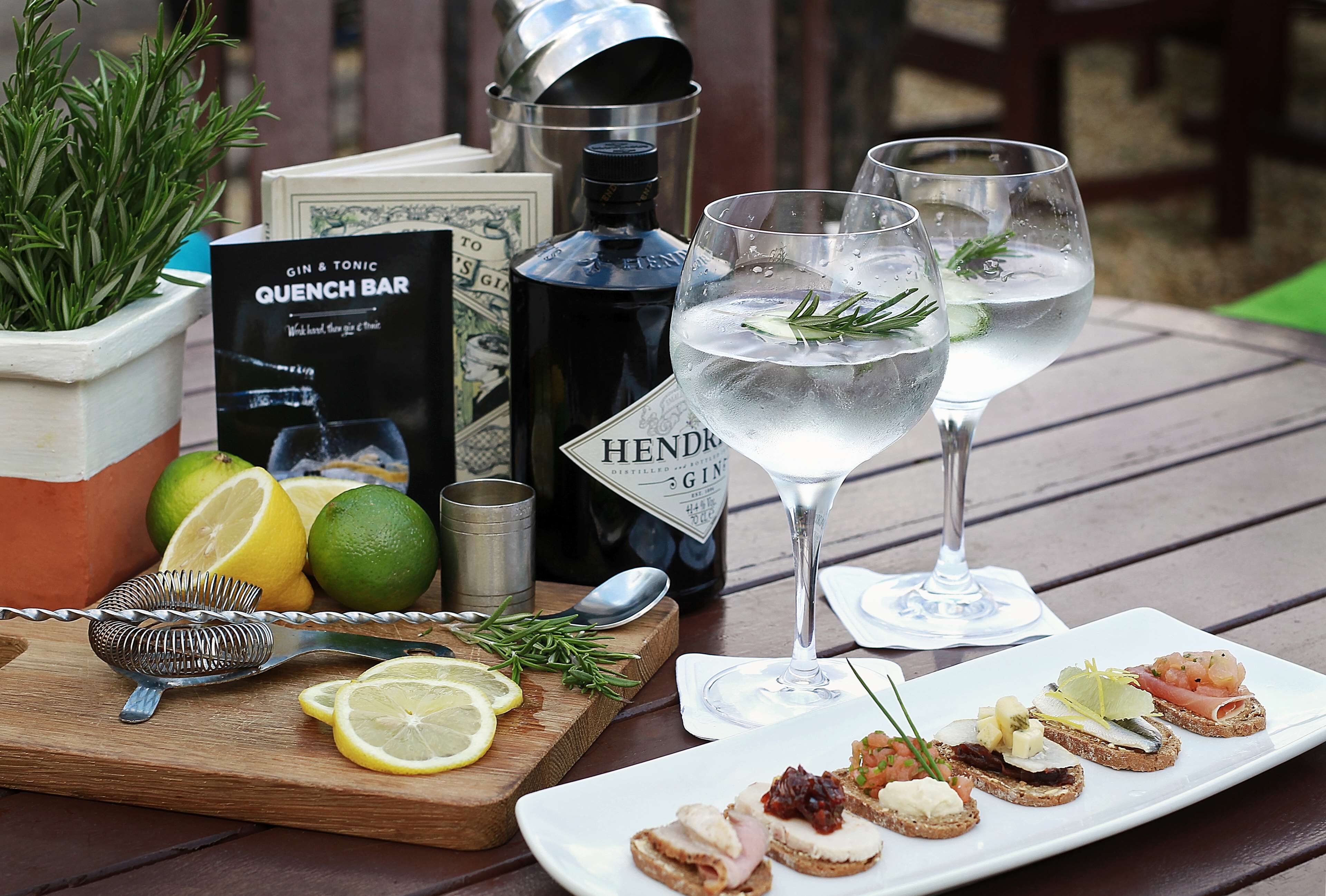 Une table avec une bouteille de gin, une bouteille d’eau tonique, un menu et une assiette de hors-d’œuvre.