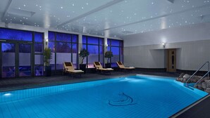 A swimming pool with a clear blue tile floor, sun loungers, potted plants, and large windows with a view of the outside.