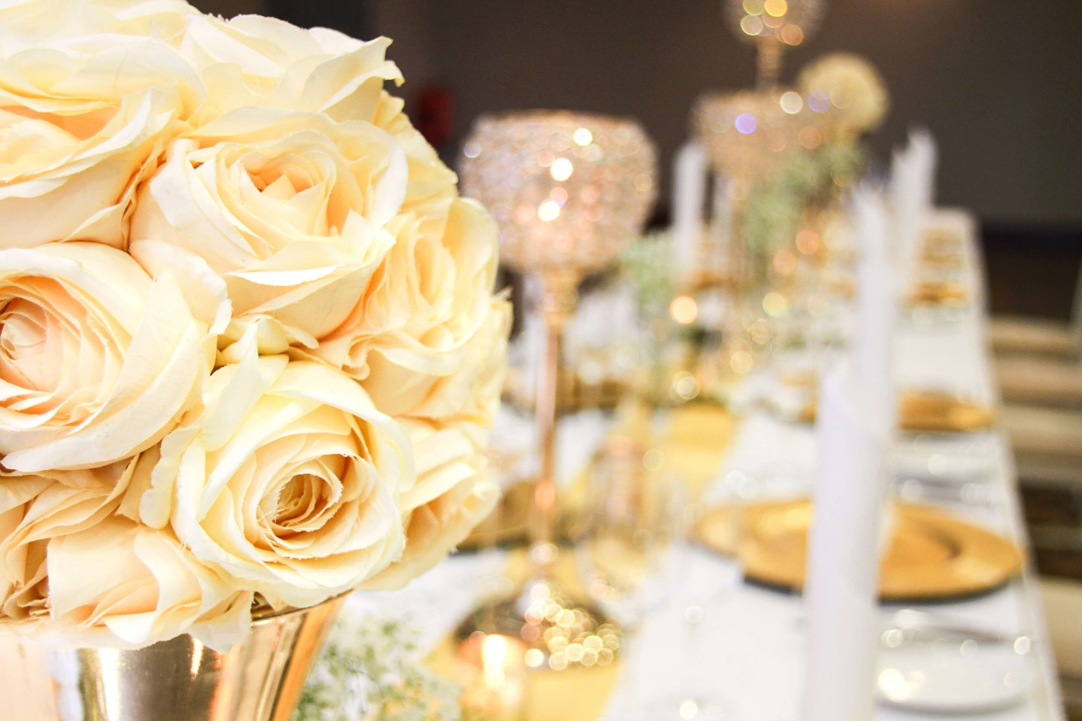 A table set with white tablecloths, gold plates, and a bouquet of pale yellow roses.