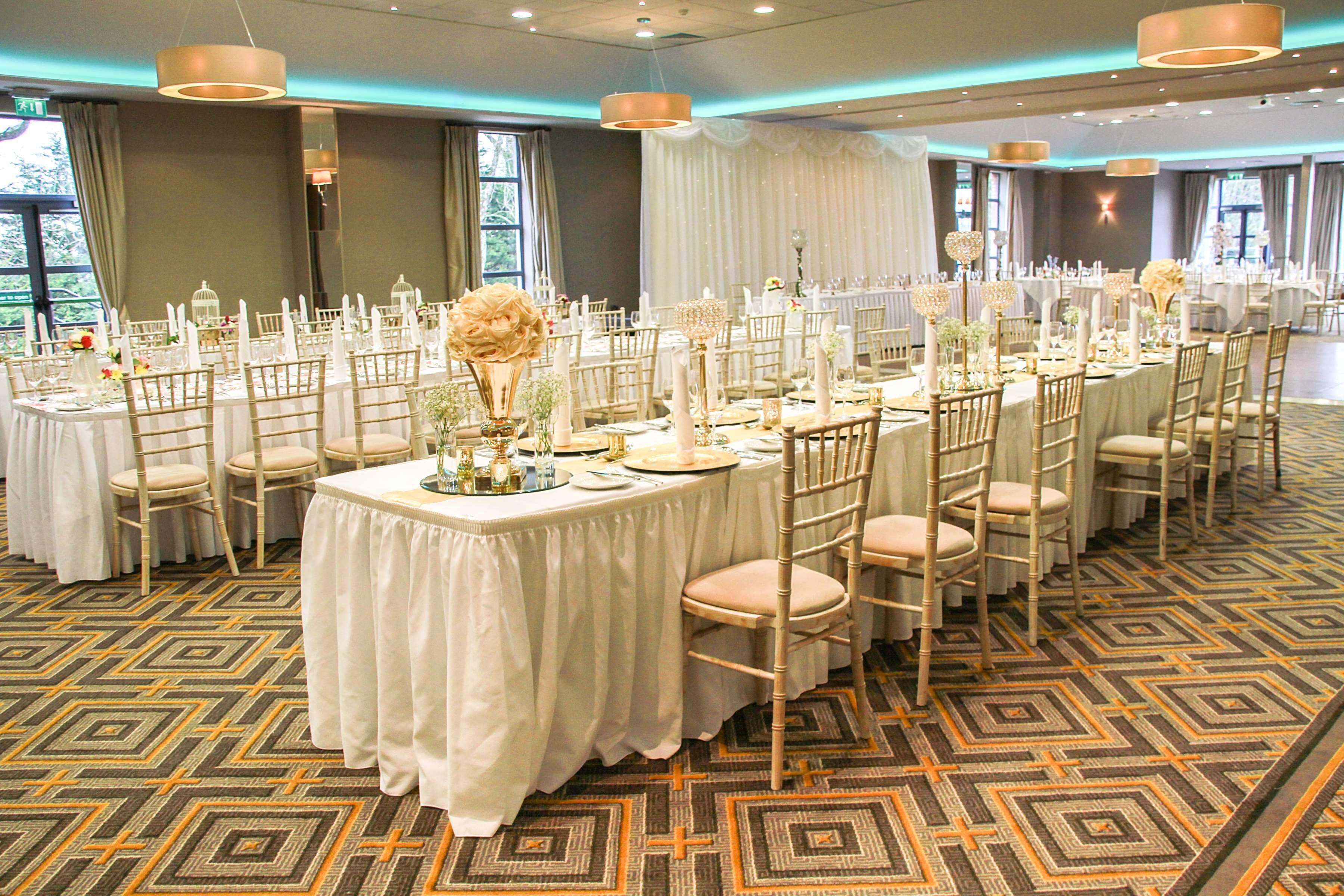 Un salón de banquetes con mesas redondas, manteles blancos y sillas doradas.