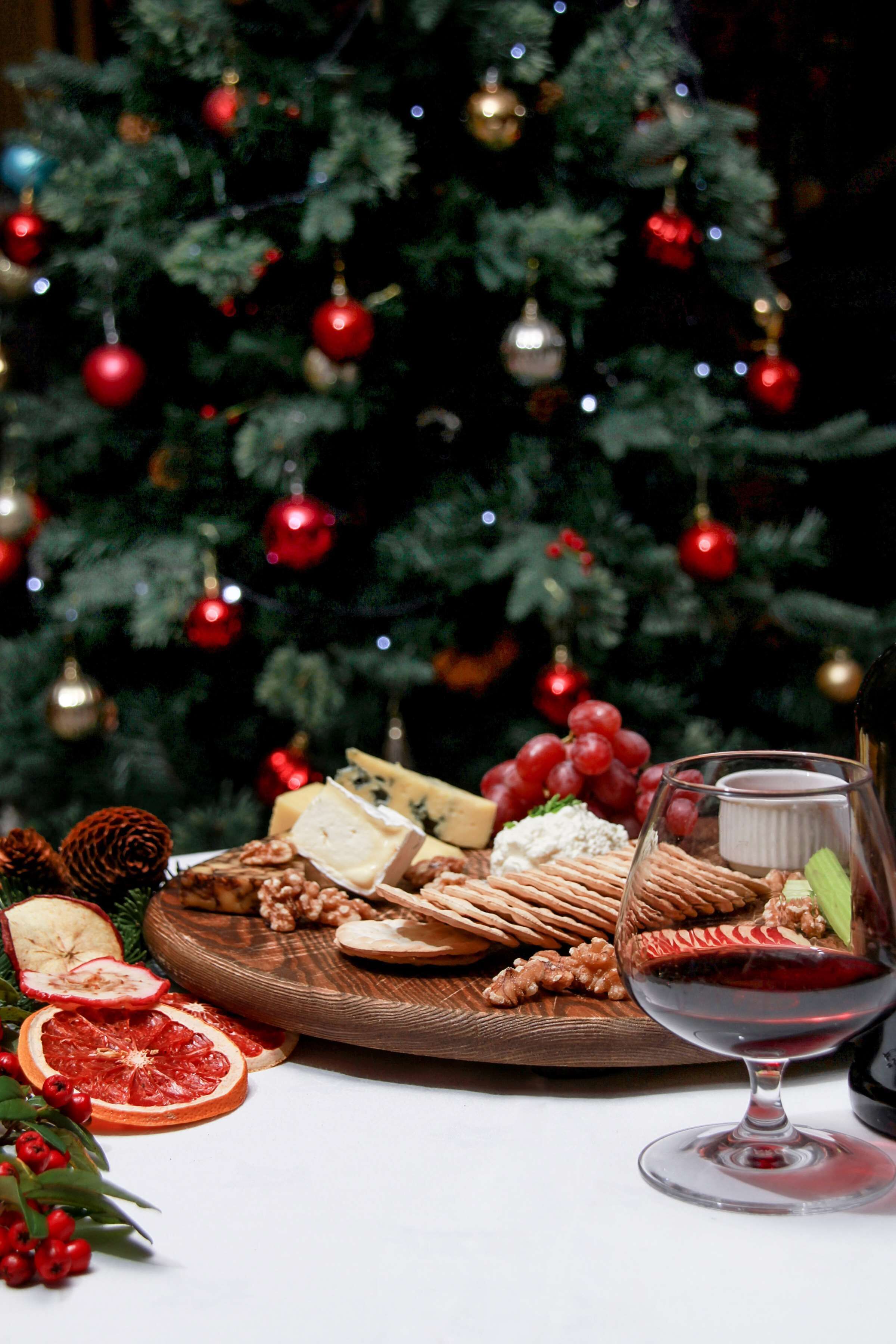 Una mesa festiva con una variedad de quesos, frutos secos y frutas, acompañada de una copa de vino tinto, frente a un árbol de Navidad decorado.