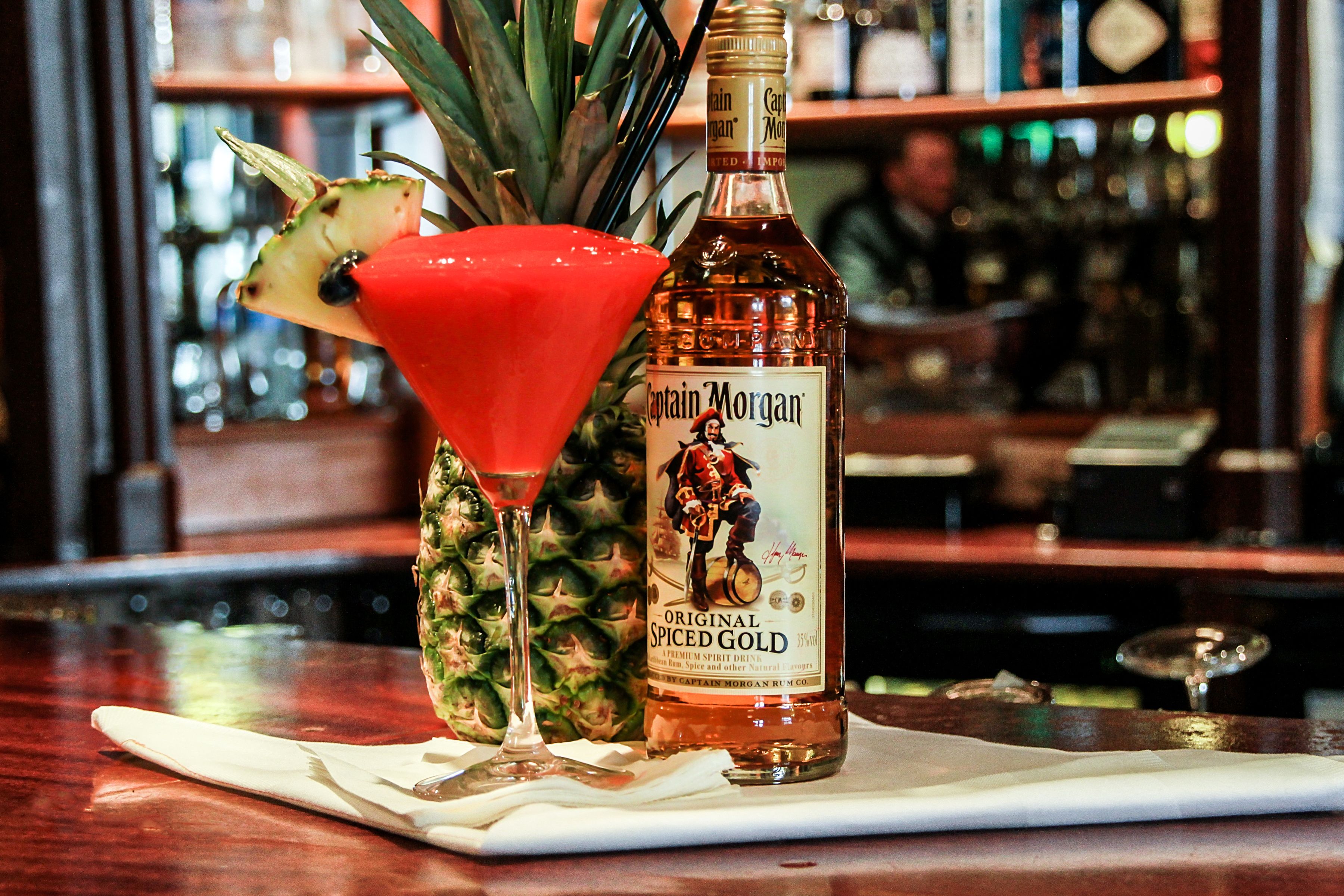 Una botella de Captain Morgan Original Spiced Gold sobre una barra con un cóctel rojo y una guarnición de piña.