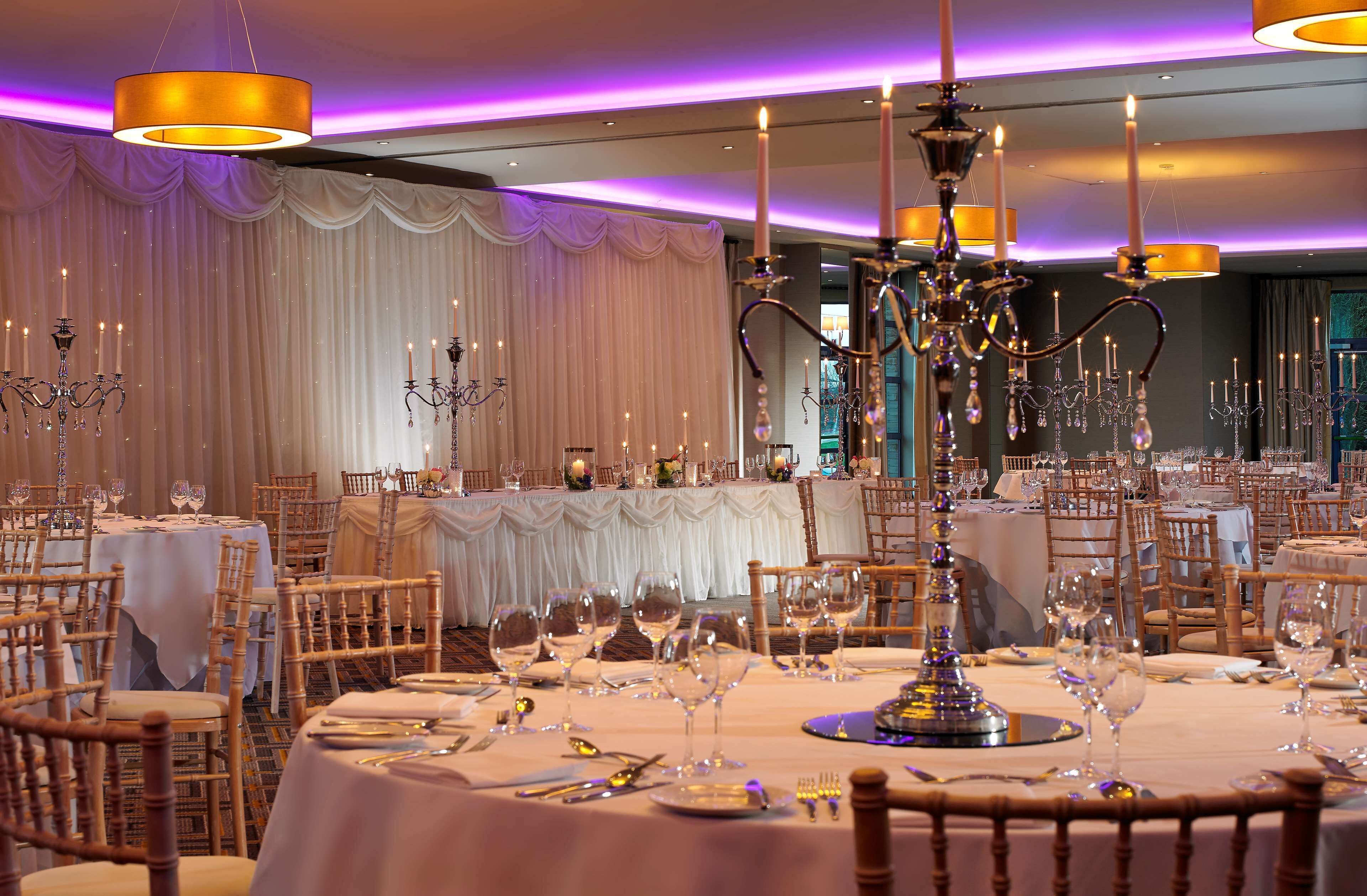 Un salón de banquetes con mesas redondas dispuestas para un evento formal, con candelabros y cortinas decorativas.