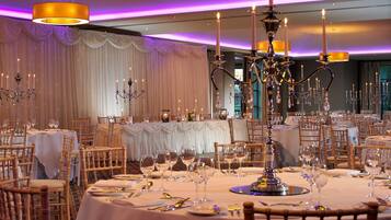 Un salón de banquetes con mesas redondas dispuestas para un evento formal, con candelabros y cortinas decorativas.