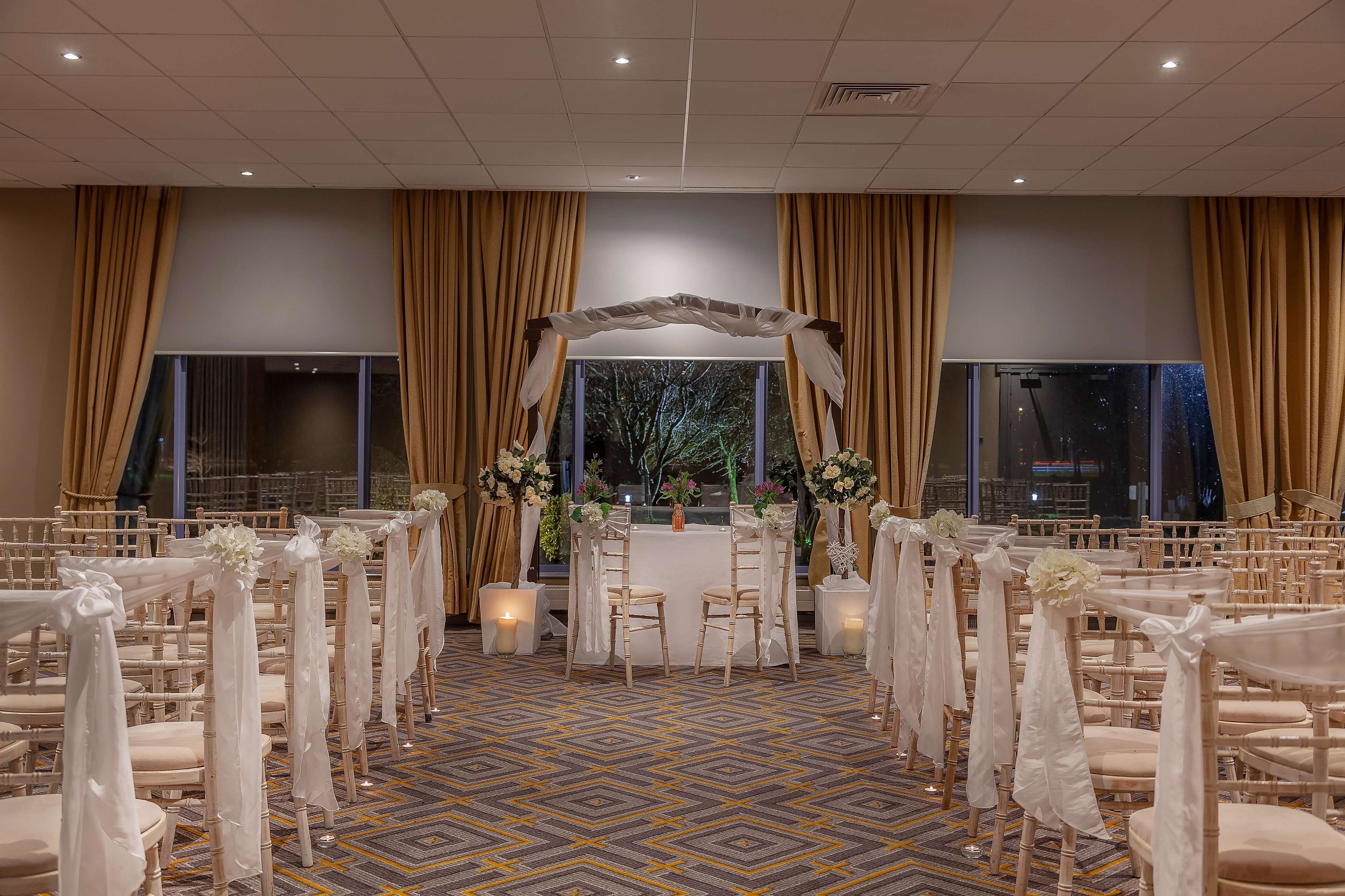 Montaje para una ceremonia de boda con filas de sillas, un altar central y grandes ventanales con cortinas.