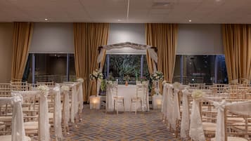 Montaje para una ceremonia de boda con filas de sillas, un altar central y grandes ventanales con cortinas.