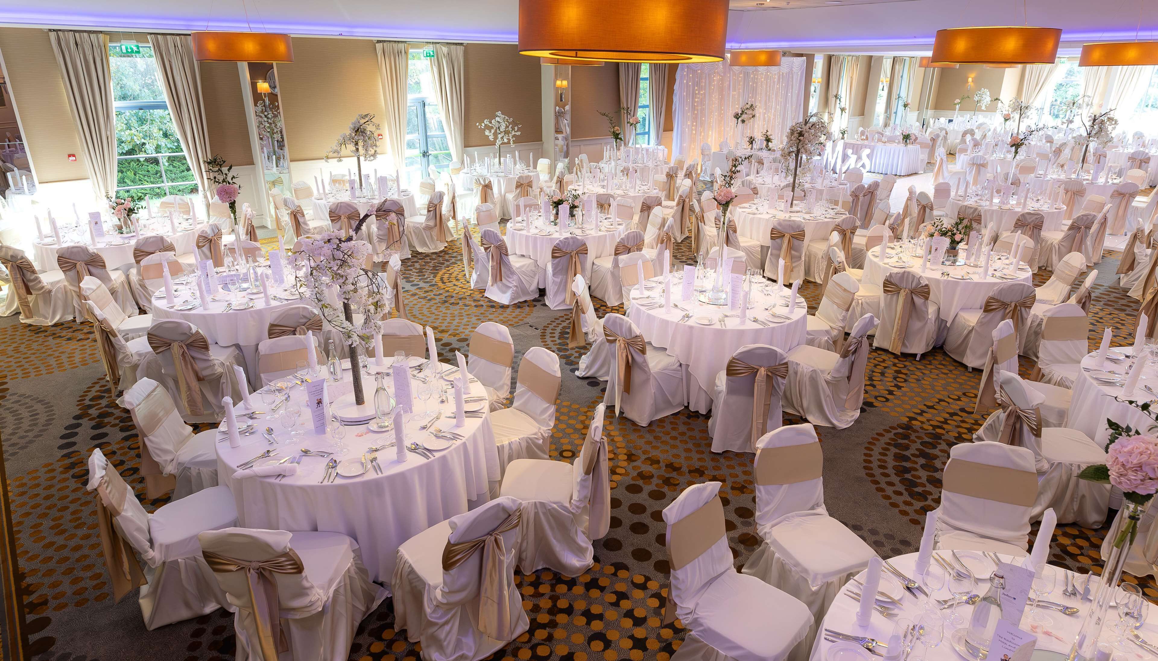 Un salón de banquetes con mesas redondas, manteles blancos y sillas adornadas con cintas doradas.