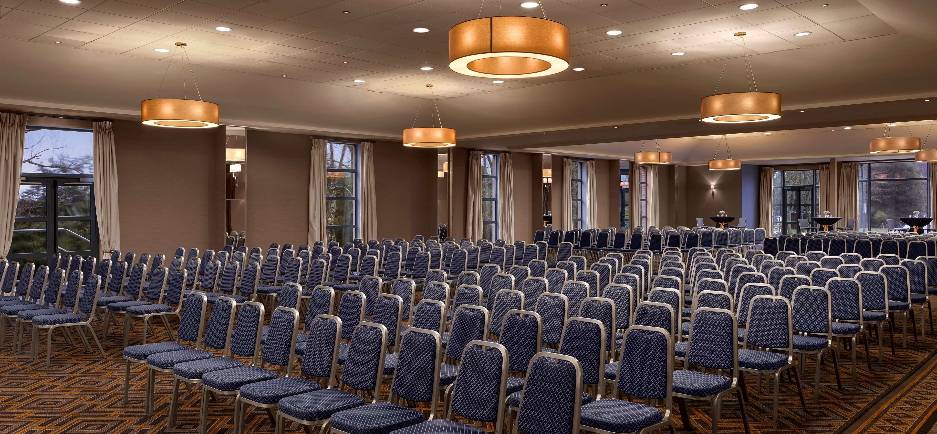 Un salón de conferencias con filas de sillas, ventanas amplias y lámparas colgantes.