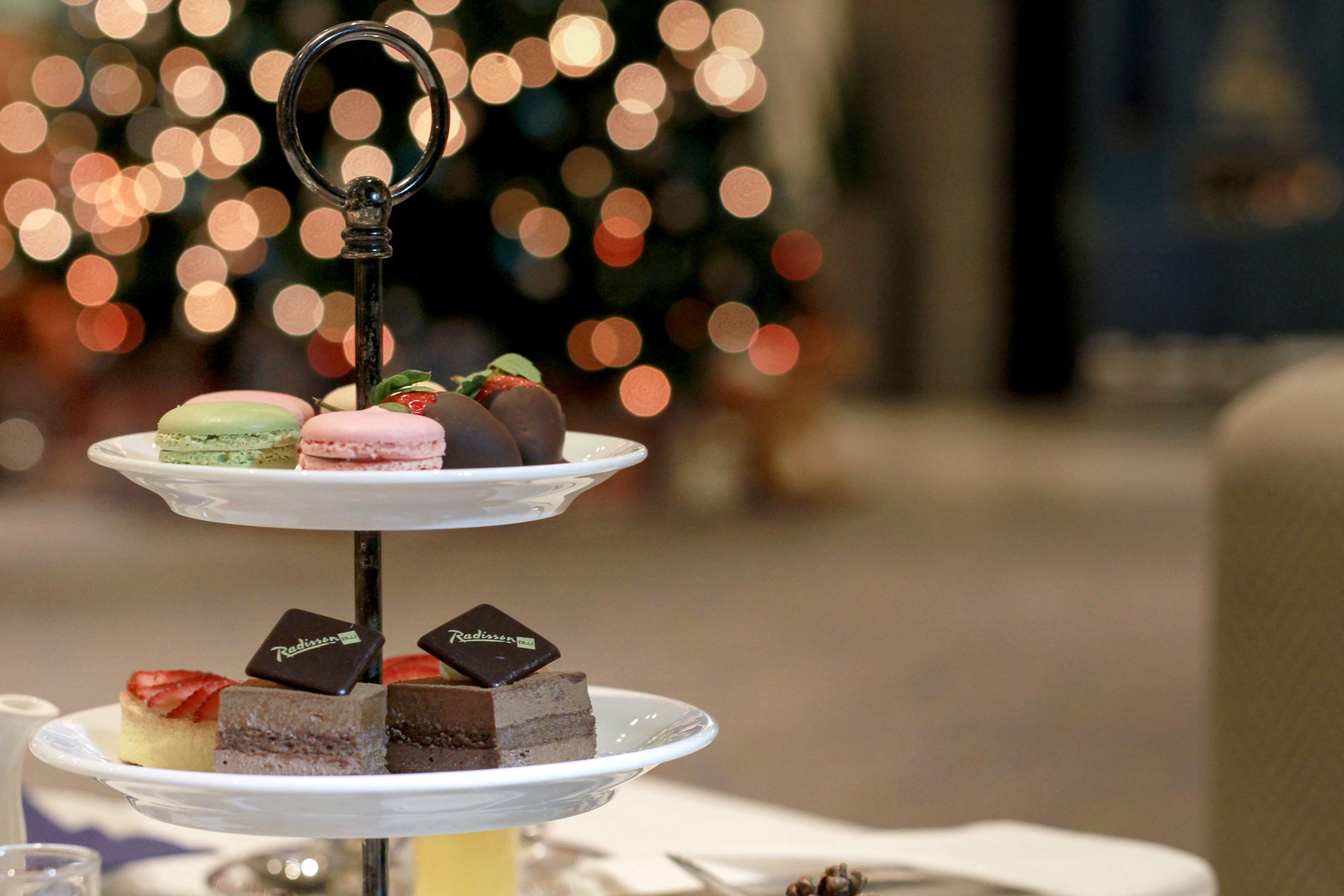 A three-tiered dessert stand with various pastries and cakes, set against a blurred background with bokeh lights.
