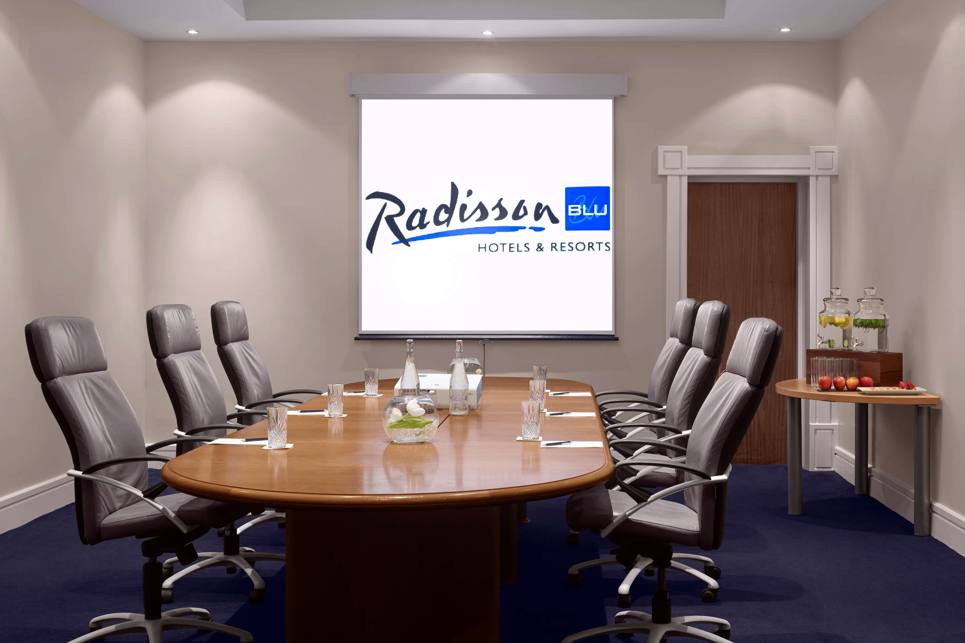 Un salón de conferencias con una gran mesa, ocho sillas y una pantalla de proyector mostrando el logotipo de Radisson BLU Hotels & Resorts.