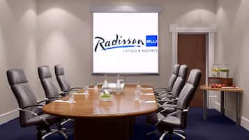 Un salón de conferencias con una gran mesa, ocho sillas y una pantalla de proyector mostrando el logotipo de Radisson BLU Hotels & Resorts.