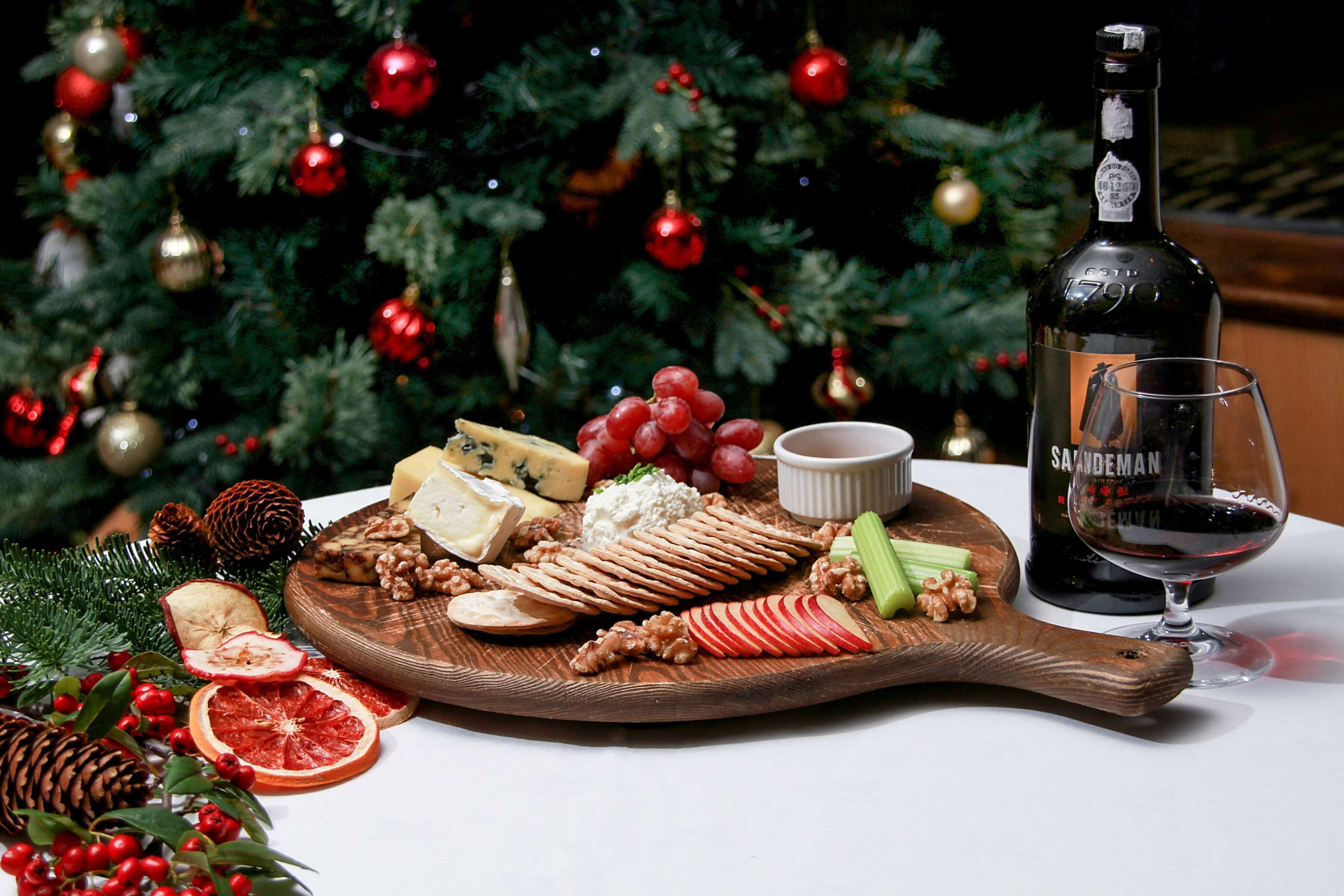 A wooden board with various cheeses, grapes, nuts, and sliced apples, accompanied by a bottle of wine and a glass, set against a Christmas tree.