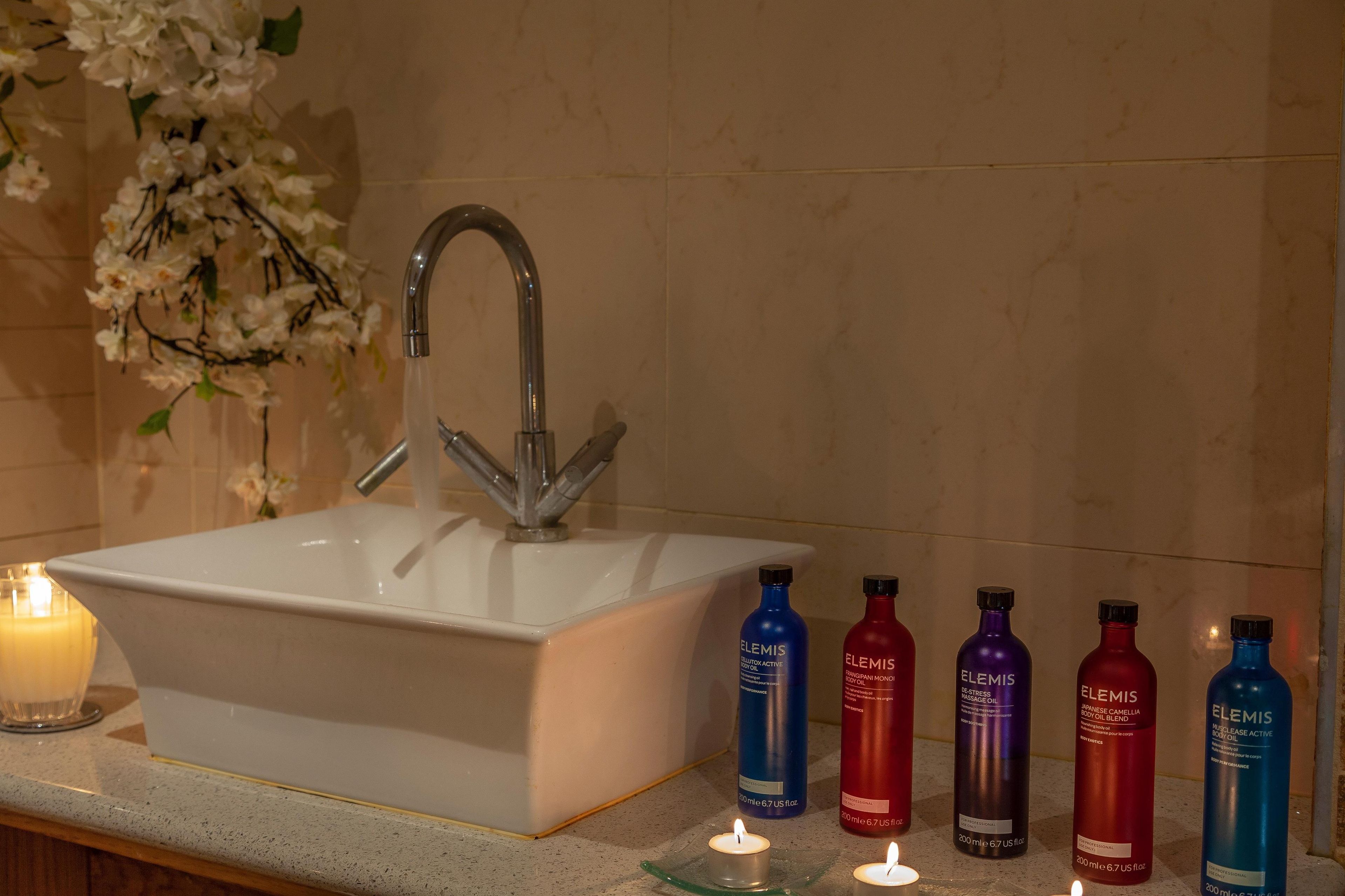 A bathroom sink with a mirror, a white candle, and a row of Elemis skincare bottles.