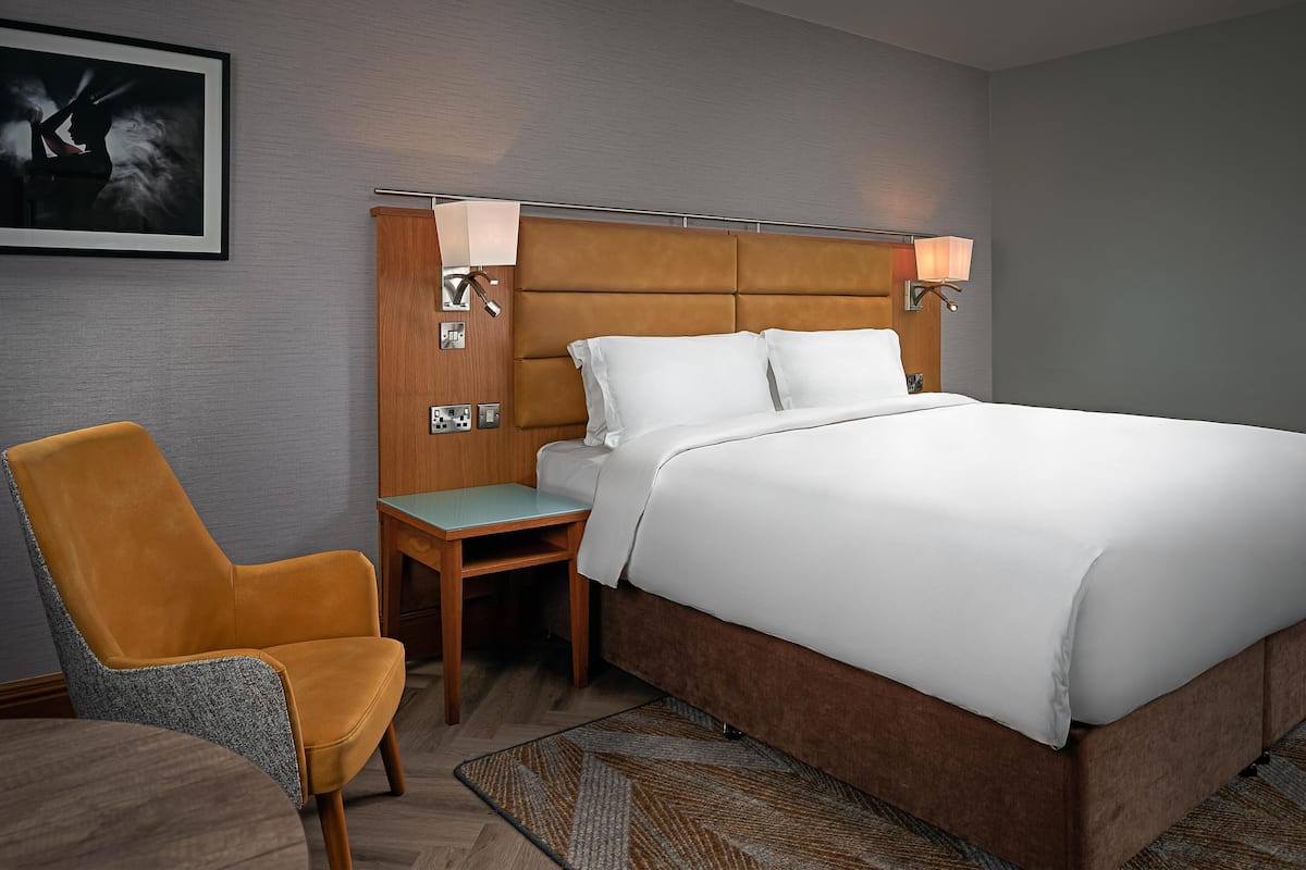 A hotel room with a bed, a chair, a nightstand, and a wall-mounted lamp.