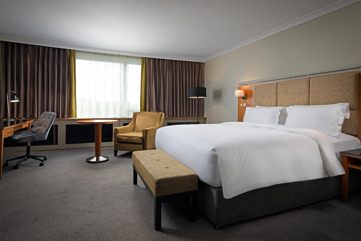 A hotel room with a large bed, a desk with a chair, a small table, and a bench.