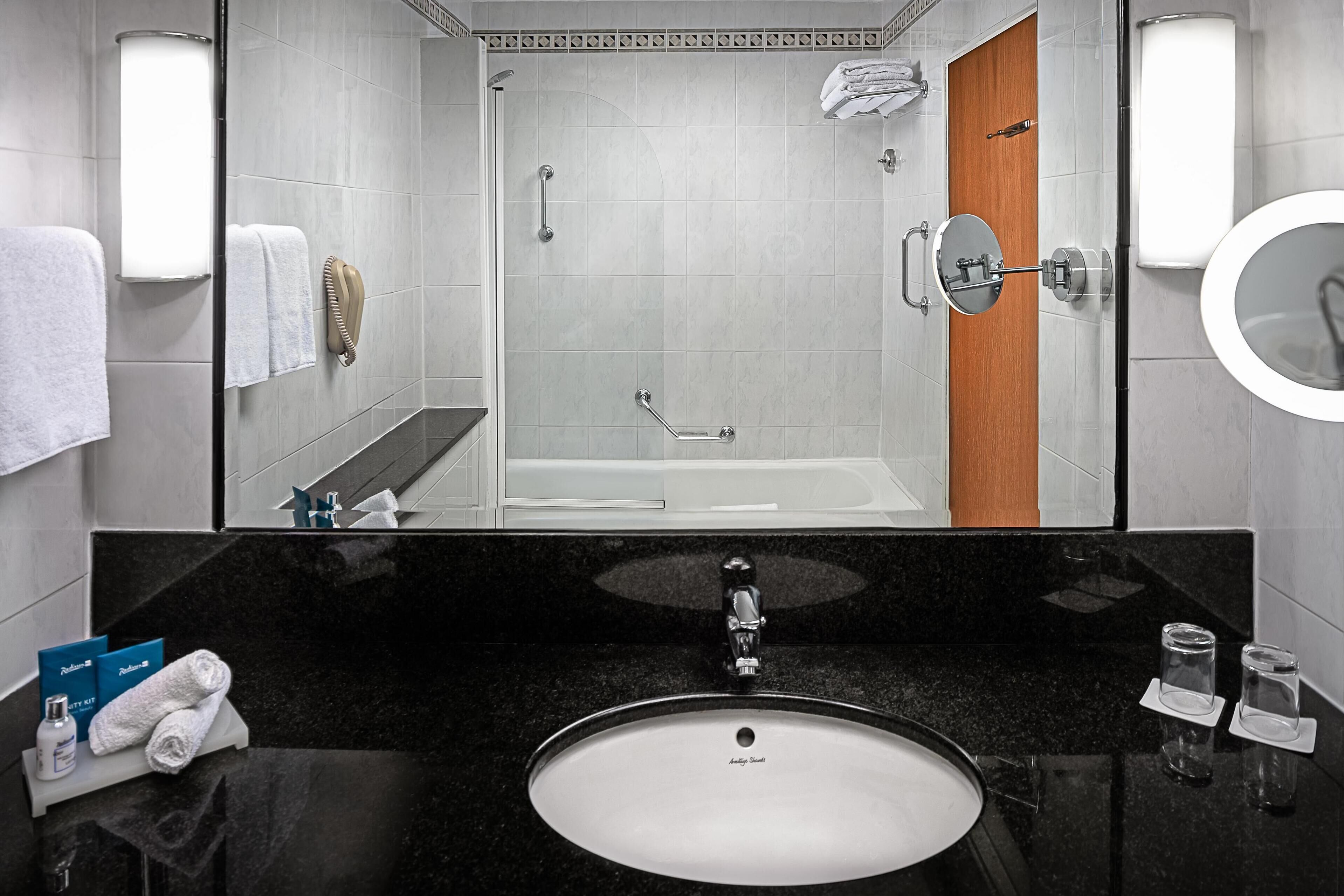 A modern bathroom with a black granite countertop, a white sink, a glass shower enclosure, and a wooden door.