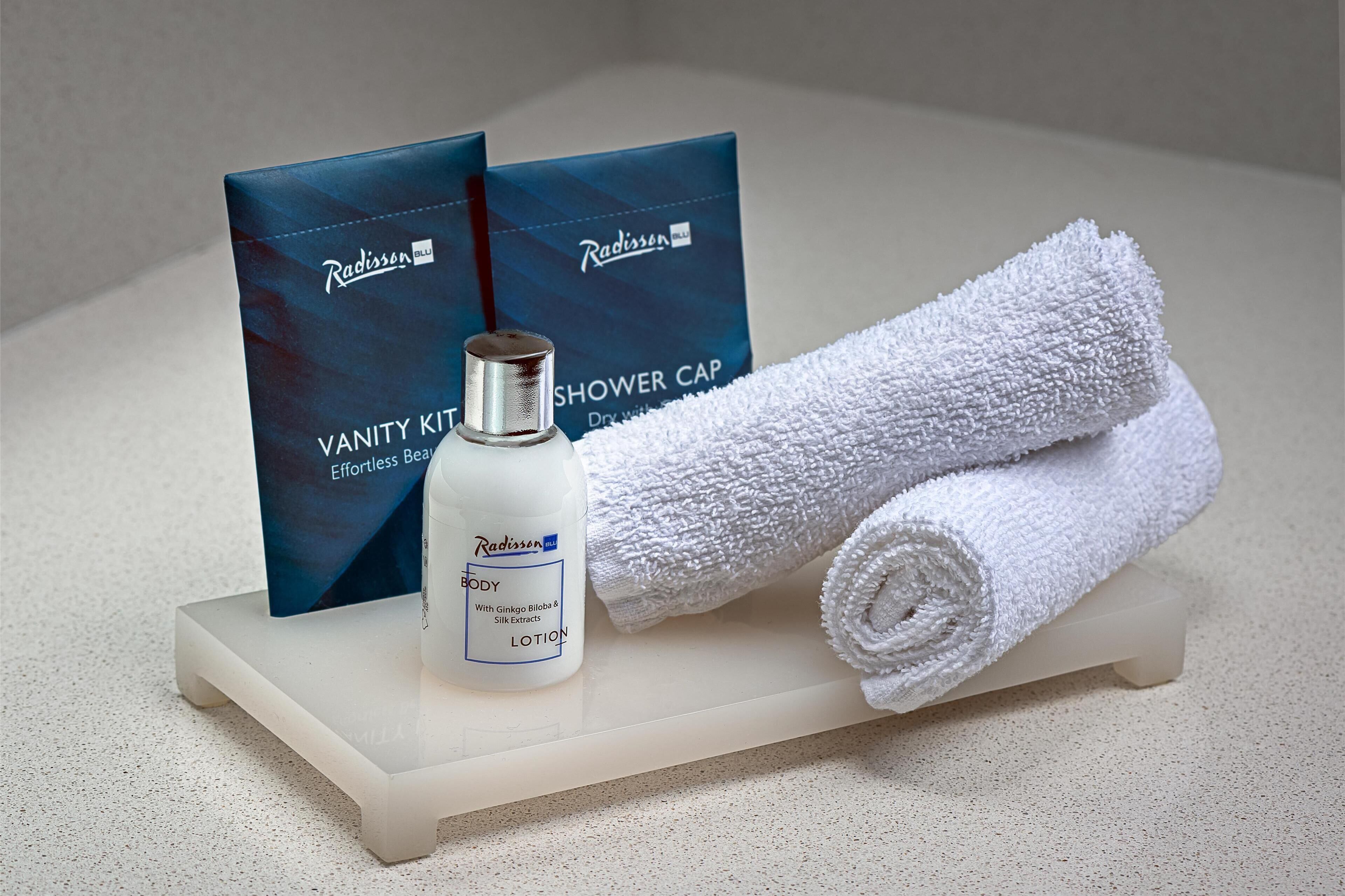 A bathroom vanity with a white towel, a bottle of Radisson body lotion, and two blue shower cap boxes.