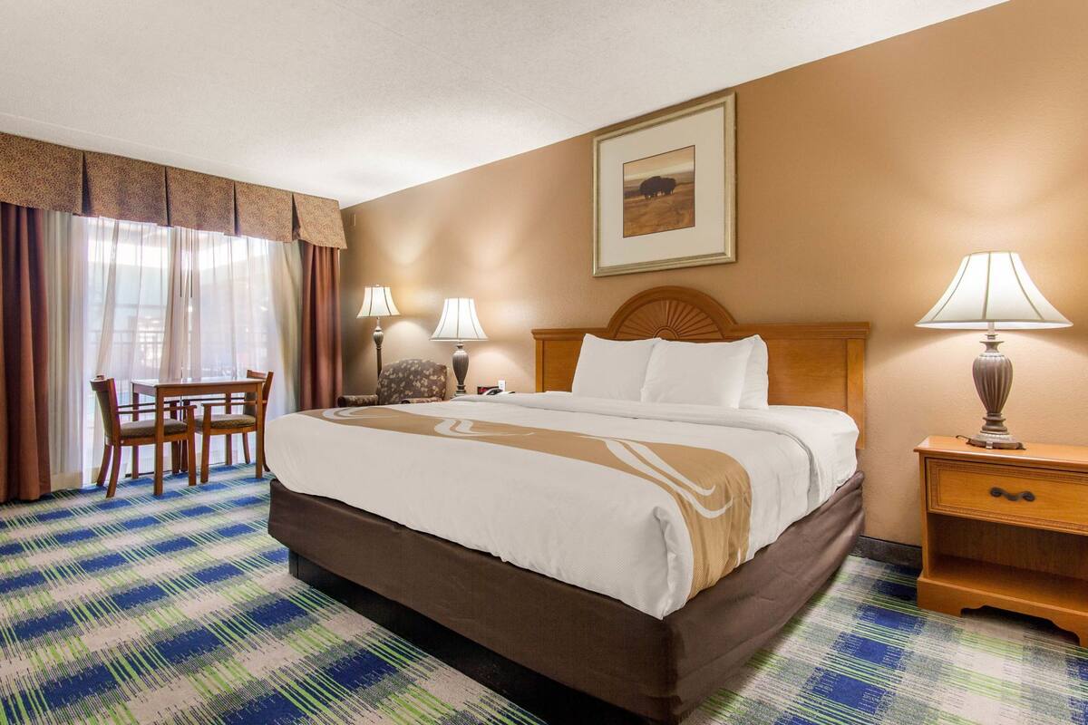 Quarto de hotel com uma cama grande, uma escrivaninha com duas cadeiras, uma mesinha de cabeceira com abajur e um quadro na parede.