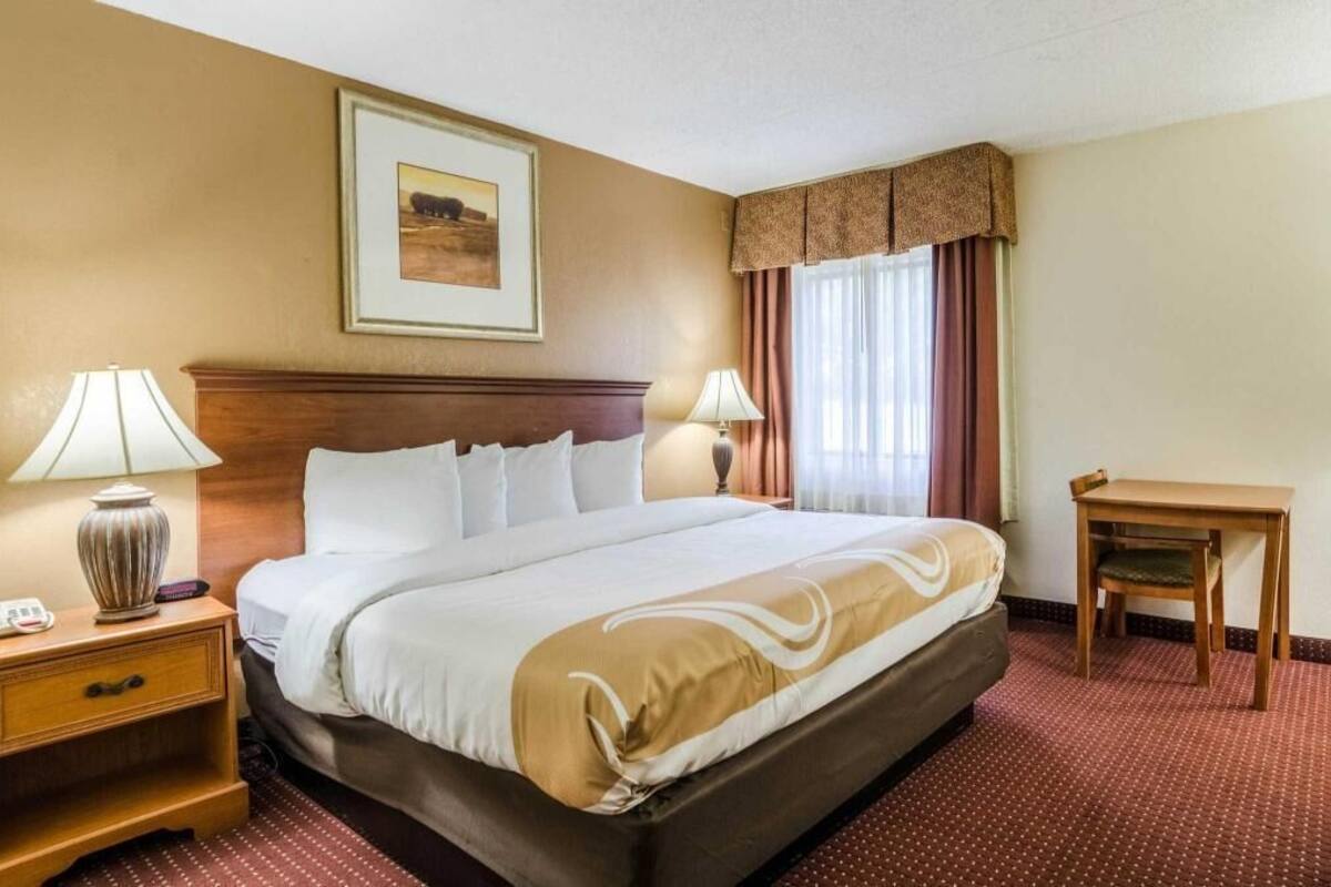 Quarto de hotel com uma cama, duas mesas de cabeceira com luminárias, uma pequena mesa de madeira e um quadro emoldurado na parede.