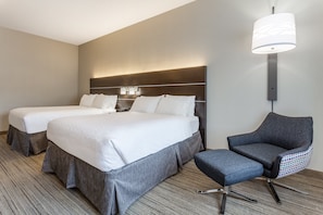 Ein Hotelzimmer mit zwei Betten, einem grauen Sessel, einem kleinen Tisch und einer Wandlampe.