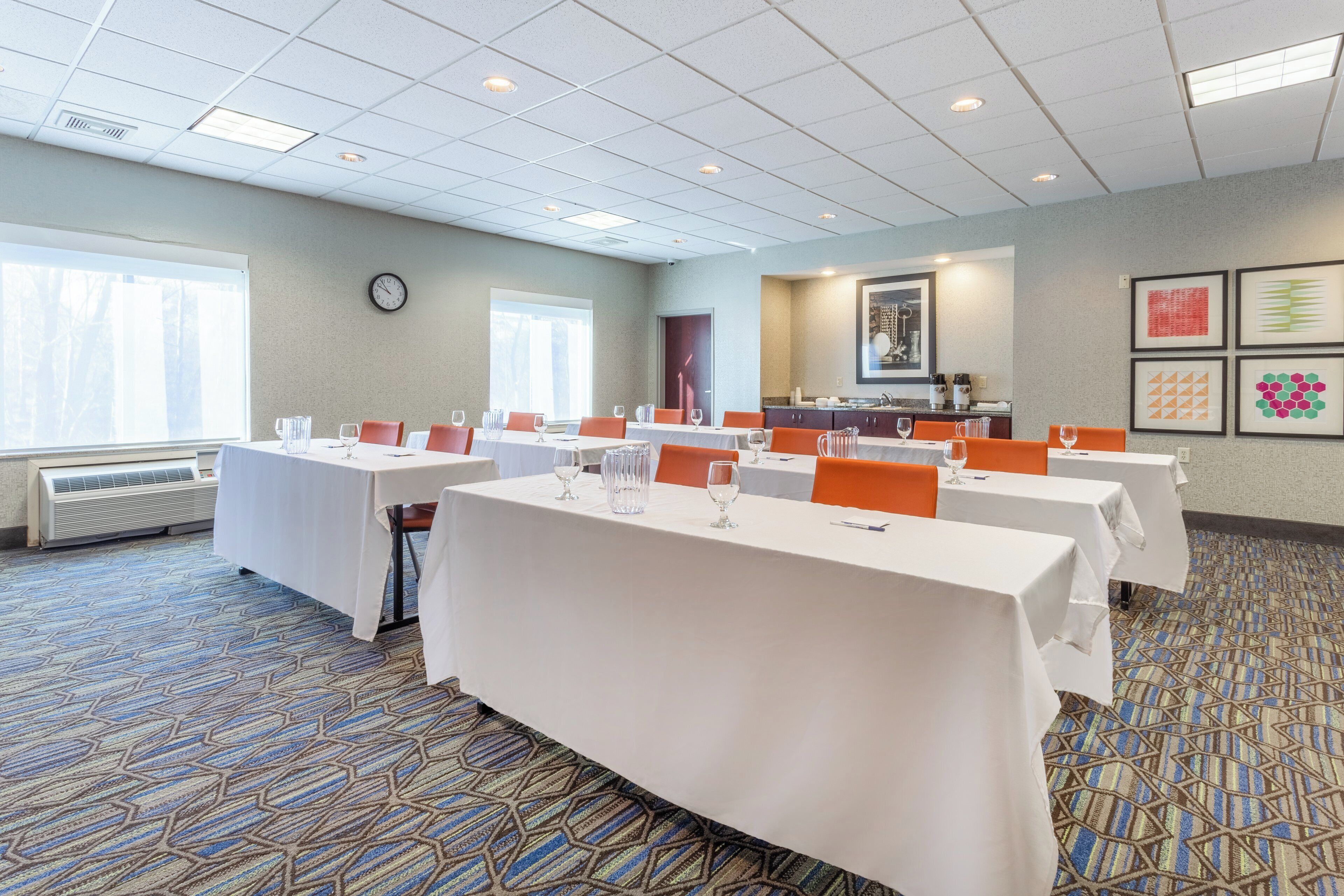 Une salle de conférence équipée de plusieurs tables, de chaises, d’une horloge, d’une fenêtre et d’un coin cuisine.