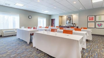 Une salle de conférence équipée de plusieurs tables, de chaises, d’une horloge, d’une fenêtre et d’un coin cuisine.