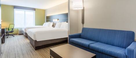 A hotel room with a bed, a blue sofa, a coffee table, a desk with a chair, and a lamp.