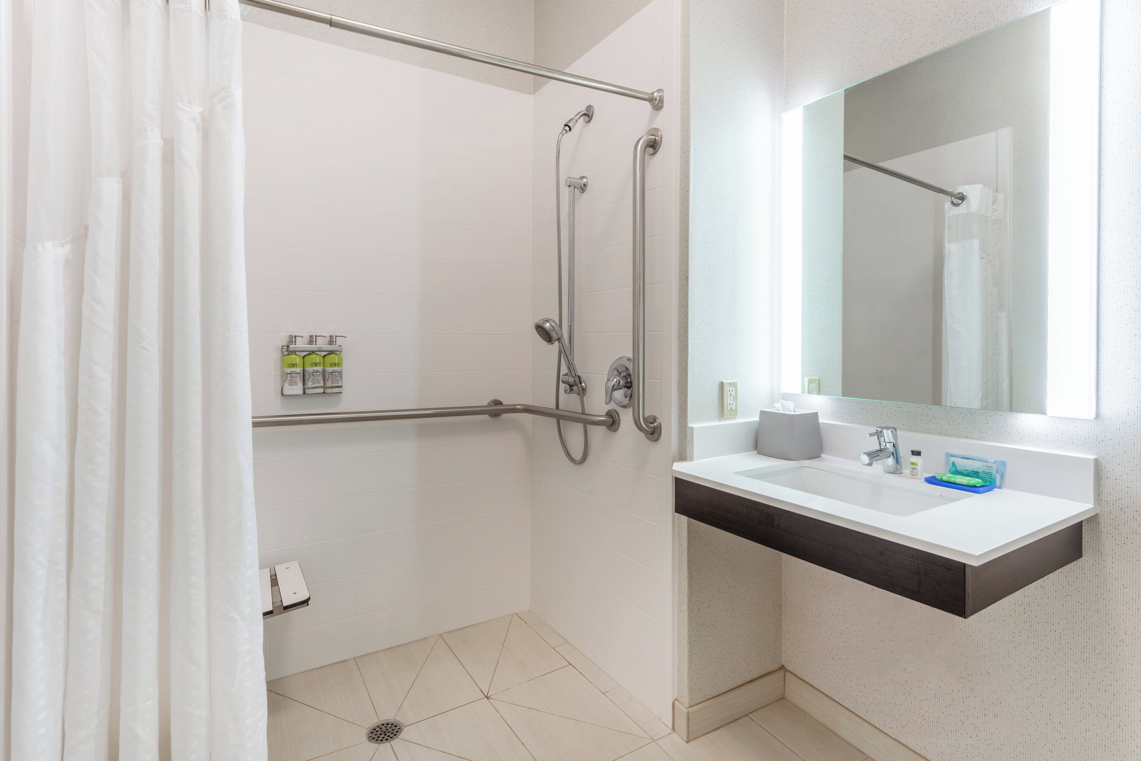 A modern bathroom with a walk-in shower, grab bars, a white sink with a dark countertop, and a mirror above the sink.