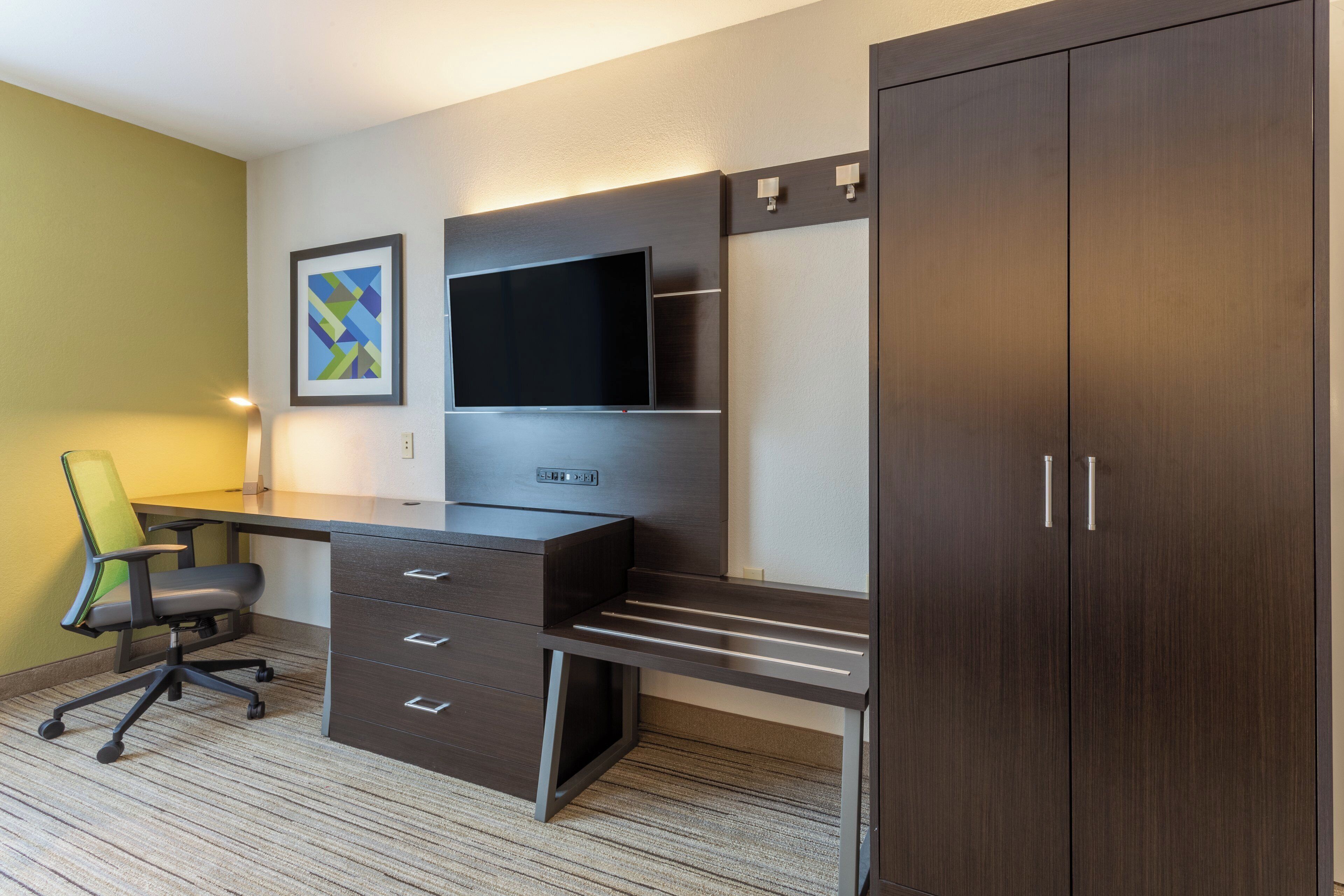 Une chambre d’hôtel avec un bureau en bois foncé, un téléviseur à écran plat, un fauteuil vert et une armoire.