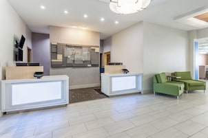 Área de recepção de hotel com dois balcões, uma kitchenette e um espaço de estar com cadeiras estofadas em verde.