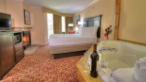 Habitación de hotel con cama, televisión, chimenea, ventana grande, baño con bañera y mesa de madera con una botella y vasos.