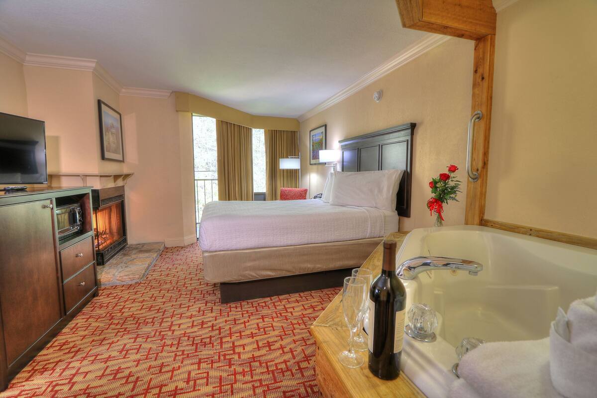 A hotel room with a bed, a TV, a fireplace, a large window, a bathroom with a bathtub, and a wooden table with a bottle and glasses.