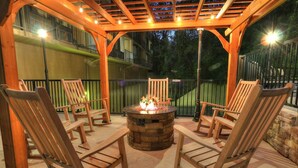 Un patio al aire libre con un hogar de piedra rodeado de sillas de madera, bajo una pérgola de madera con luces colgantes.
