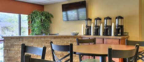 A restaurant with a stone counter, multiple coffee dispensers, a flat-screen TV, and green chairs.