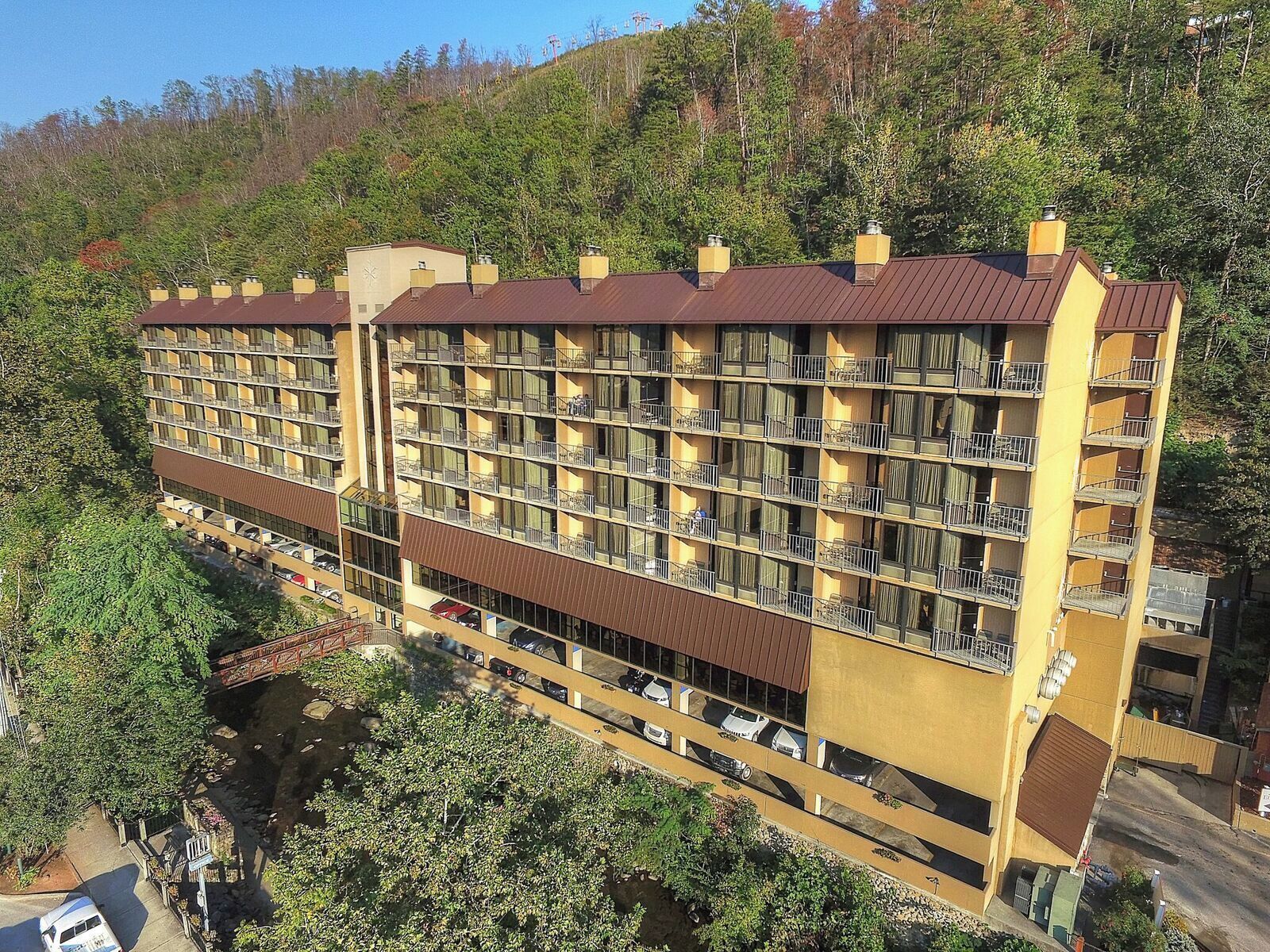 Un hotel de varios pisos con balcones, un área de estacionamiento y un paisaje montañoso de fondo.