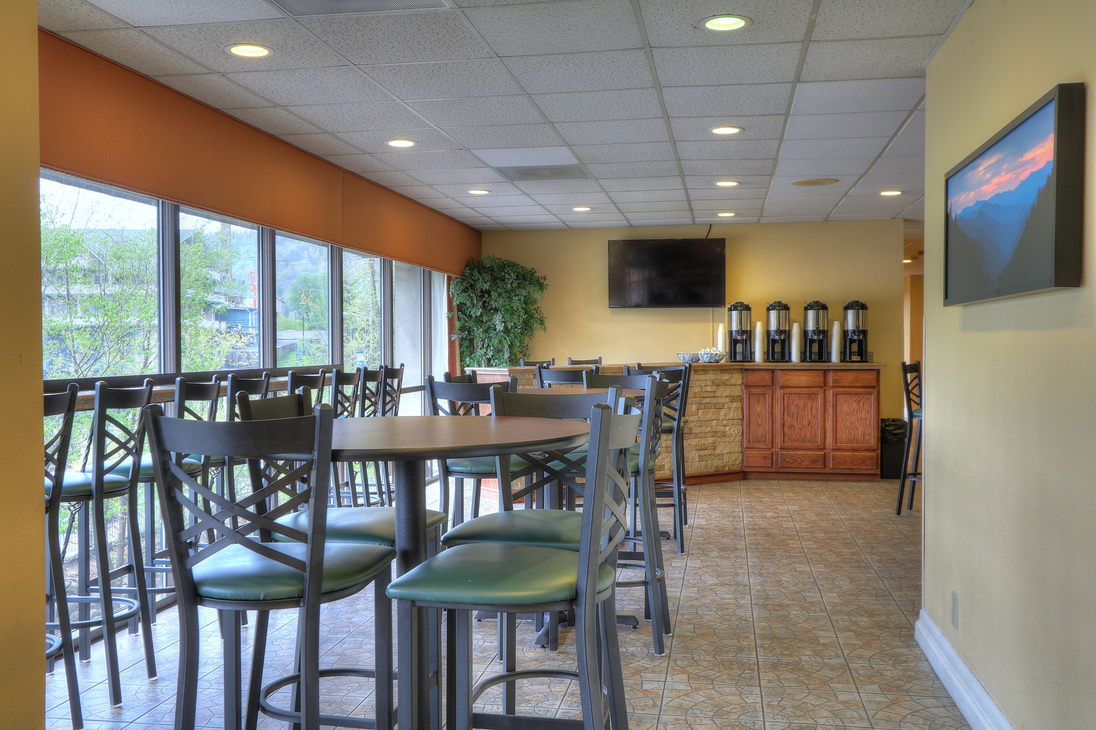 A restaurant with multiple tables and chairs, a large window with a view of trees, a television mounted on the wall, and a coffee station.