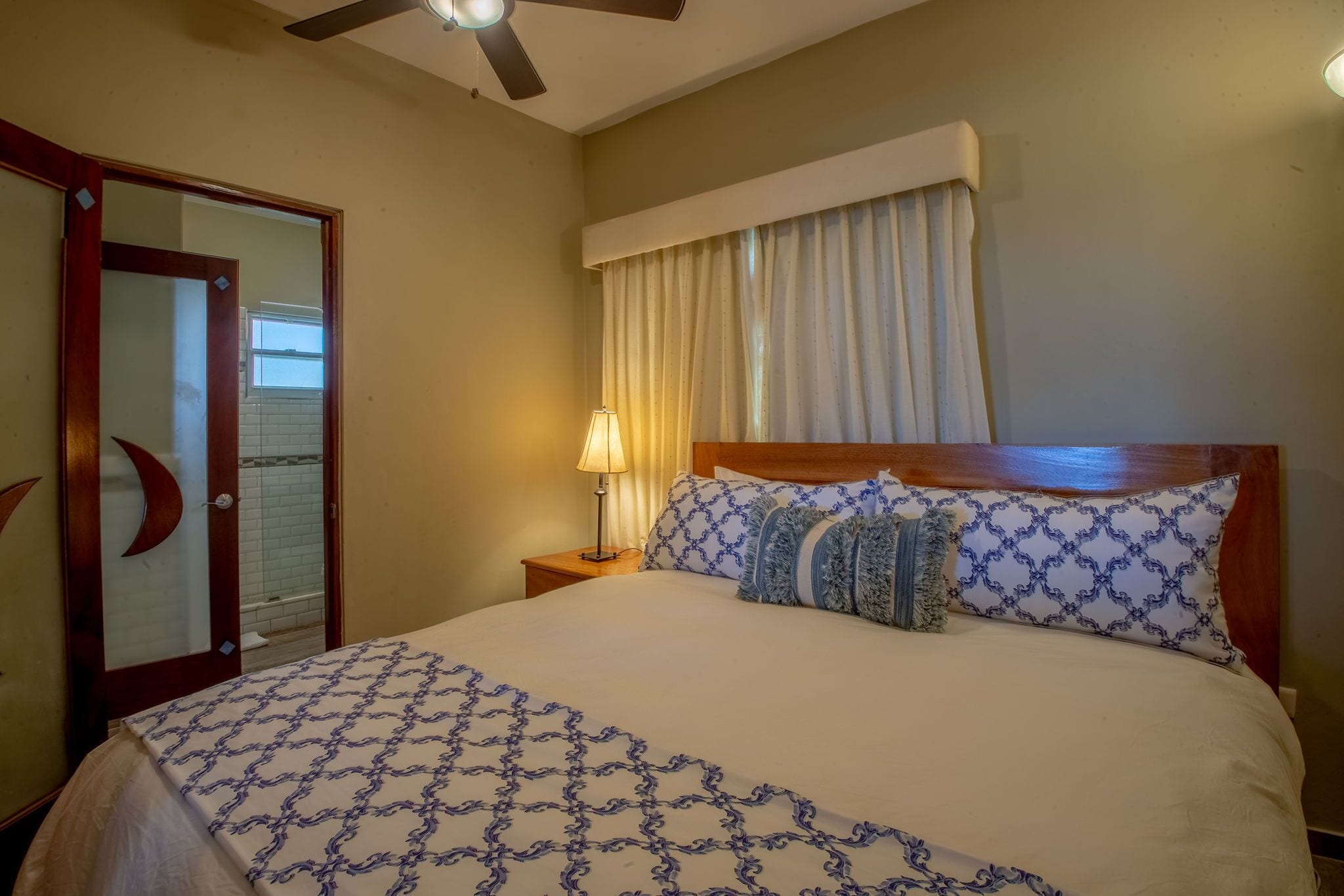 Une chambre d’hôtel avec un lit, une table de chevet, une lampe, un ventilateur de plafond et une fenêtre avec des rideaux.