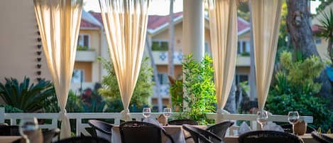 An outdoor dining area with tables and chairs, draped with white curtains, and a view of buildings and greenery in the background.