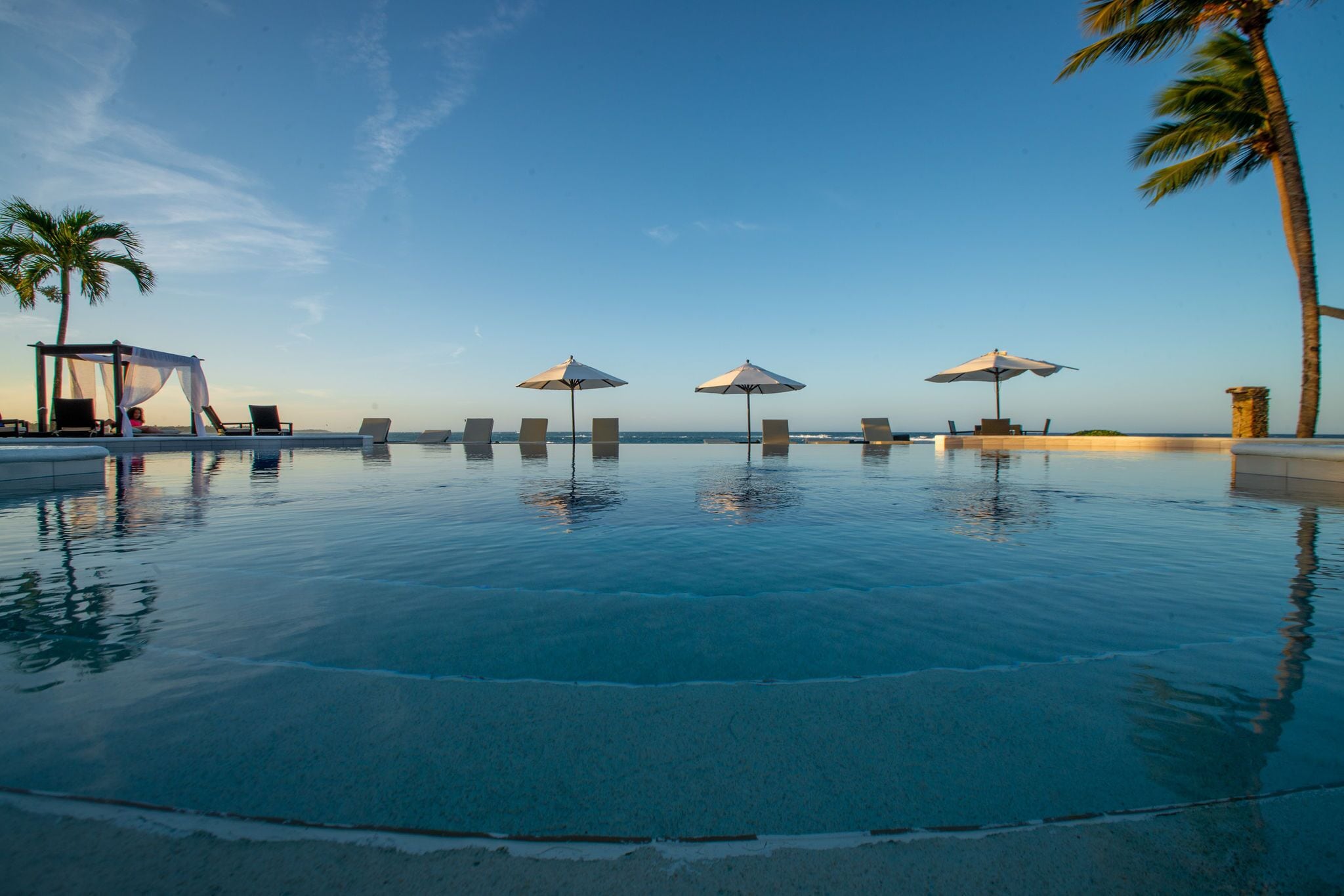 A serene infinity pool with clear water reflecting the sky, surrounded by lounge chairs and umbrellas, and palm trees in the background.