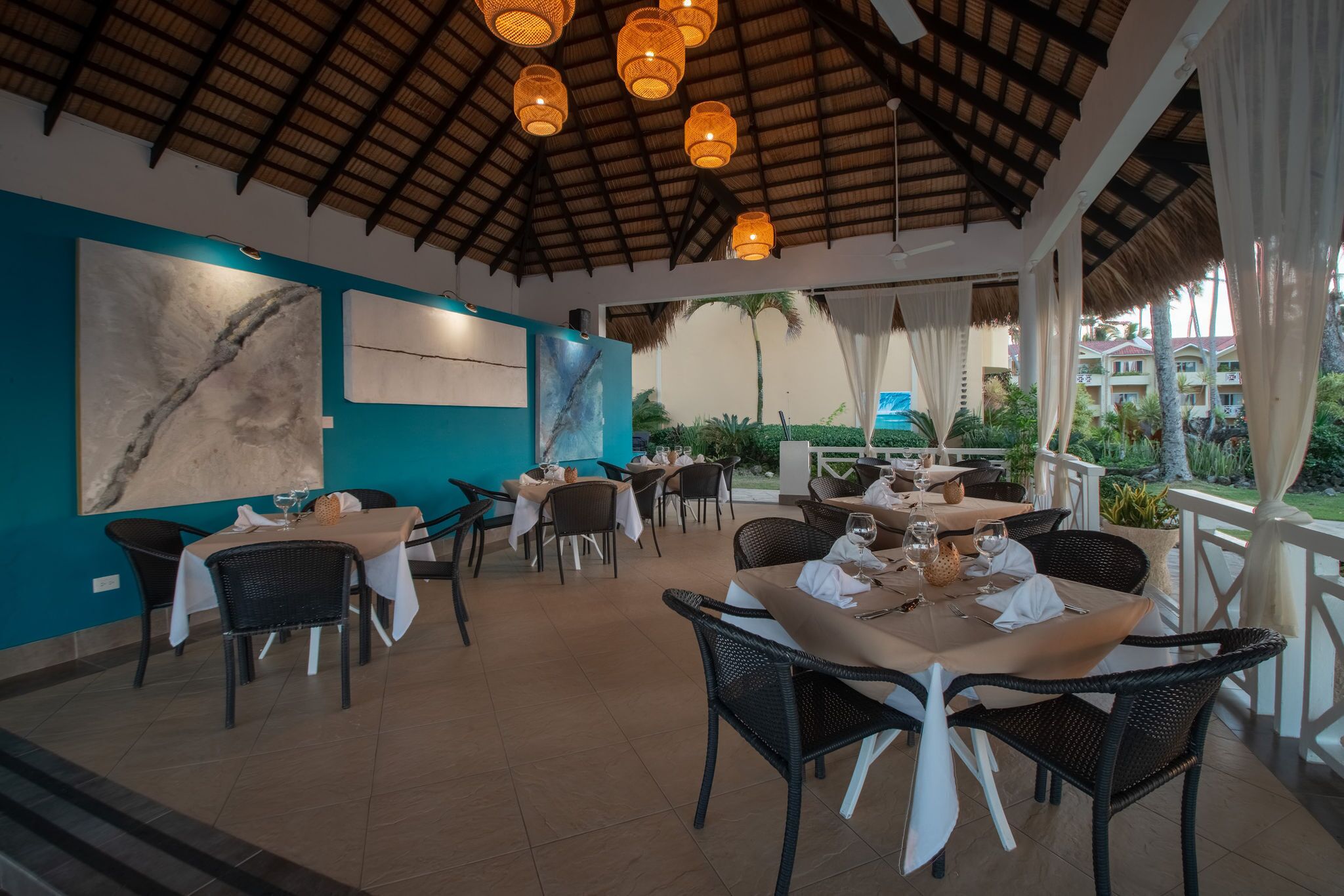 An outdoor dining area with tables and chairs, a thatched roof, and a view of a building in the background.