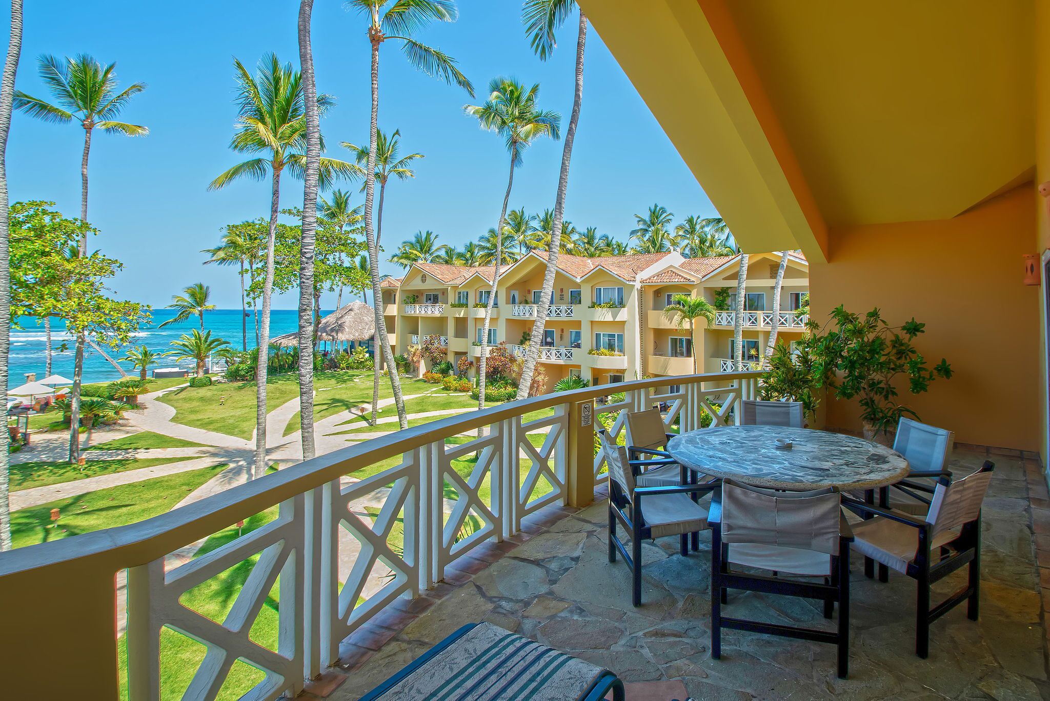 A balcony with a view of a pool and palm trees, a dining area with chairs and a table, and a building with balconies.