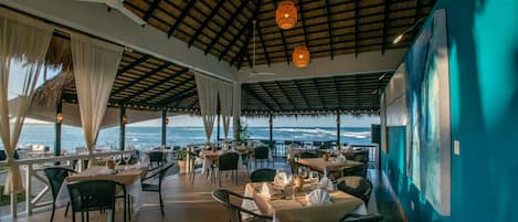 An outdoor dining area with tables and chairs, a thatched roof, and a view of the ocean.